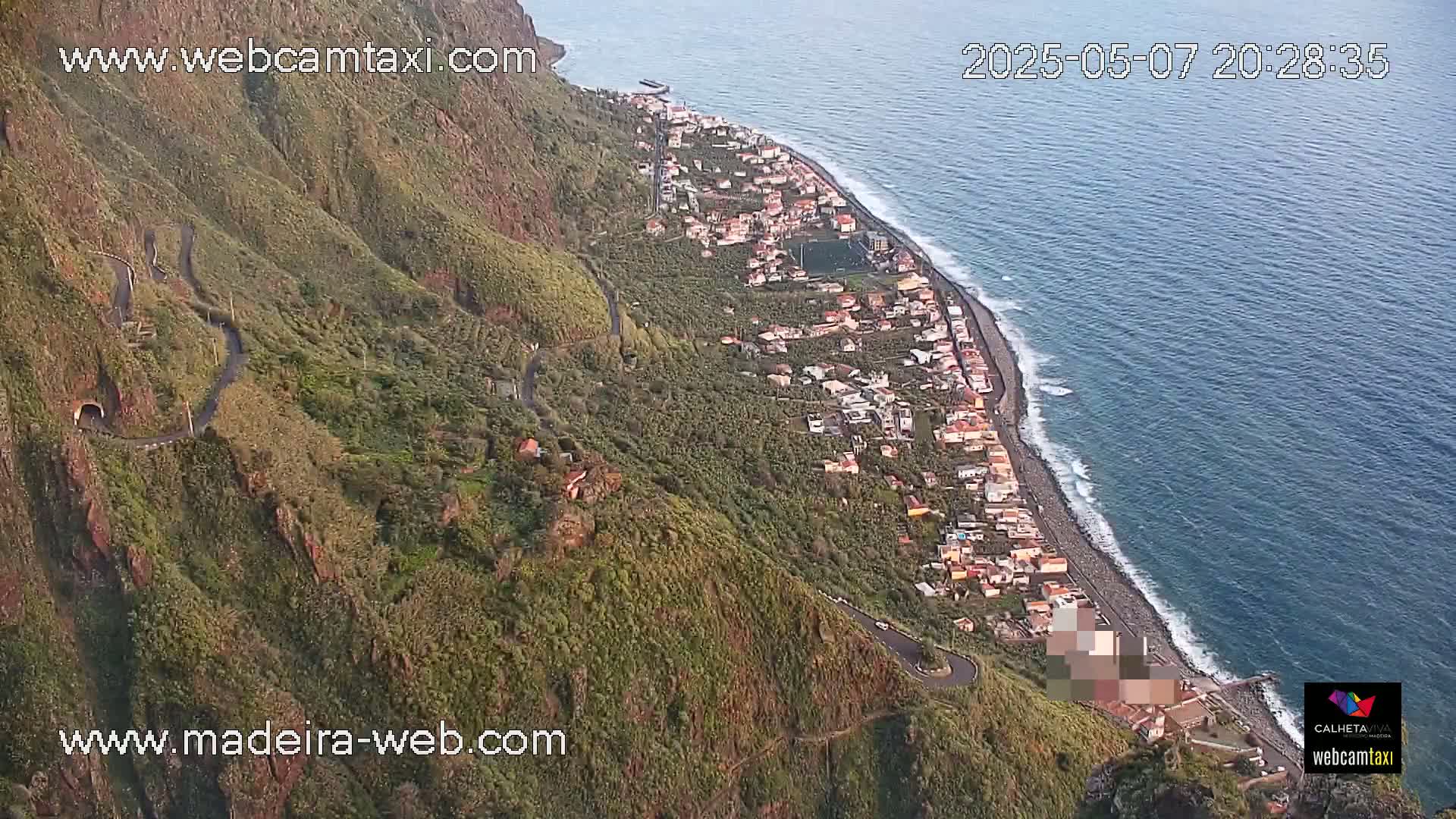 An aerial view shows a coastal town nestled between a steep, verdant mountain and a calm ocean.