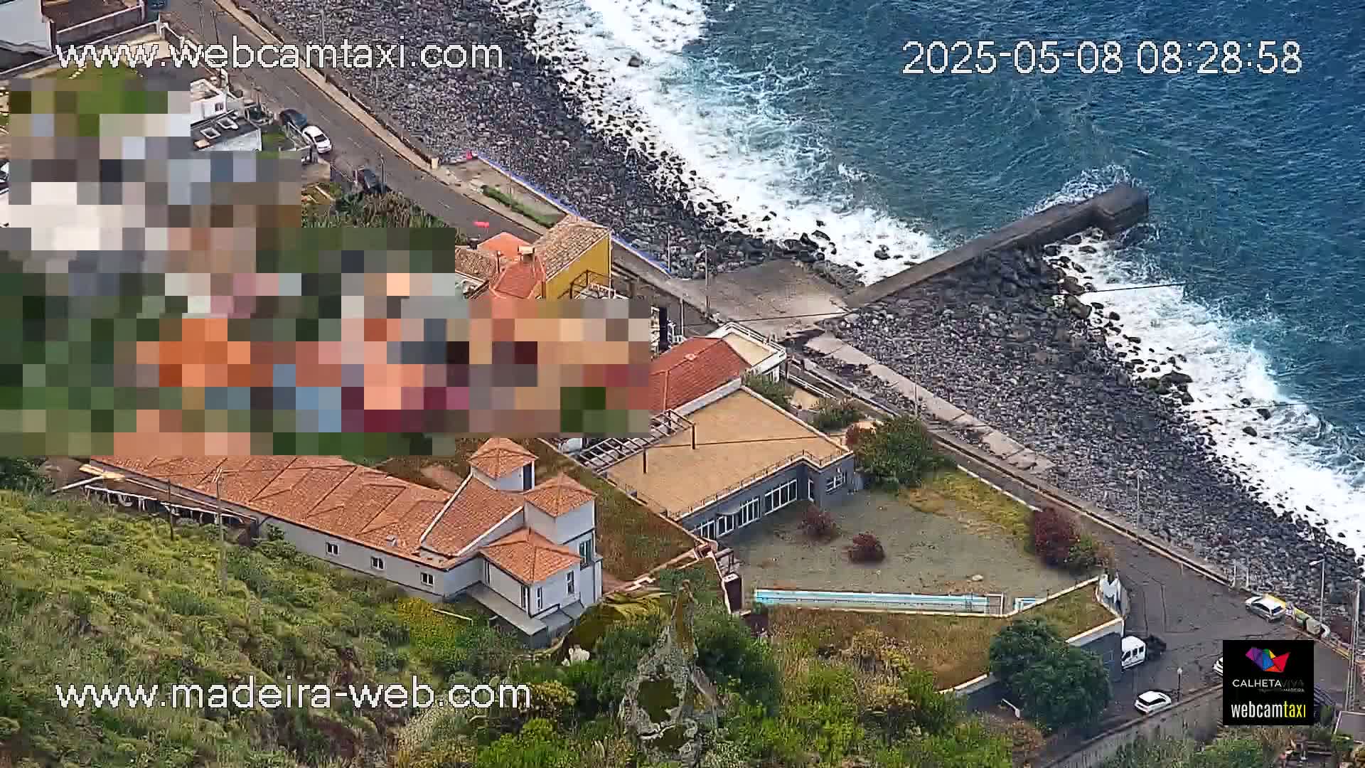 An aerial view shows a rocky coastline with crashing waves, a road, and several buildings, including a large house with a pool.