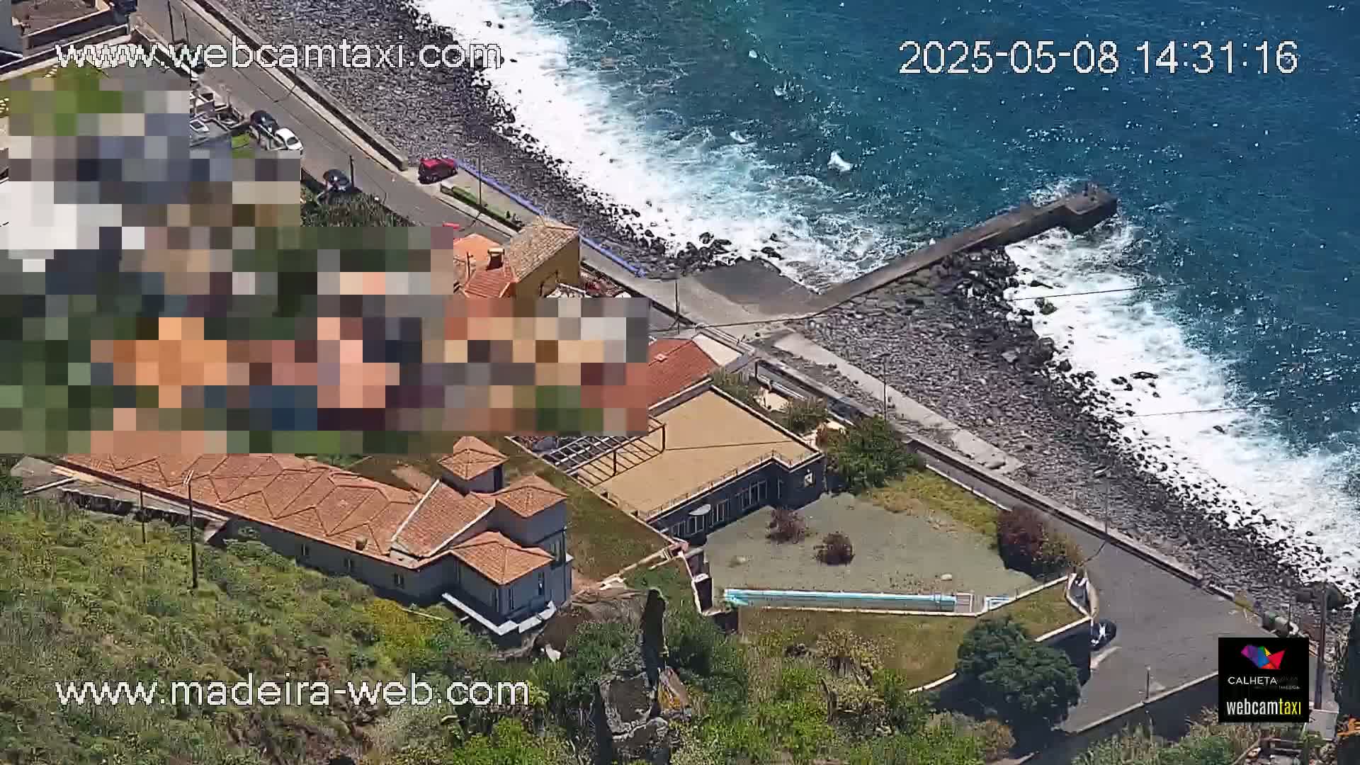 An aerial view shows a coastal property with a tiled roof and a small pool, overlooking a rocky shoreline with waves crashing against it on a sunny day.