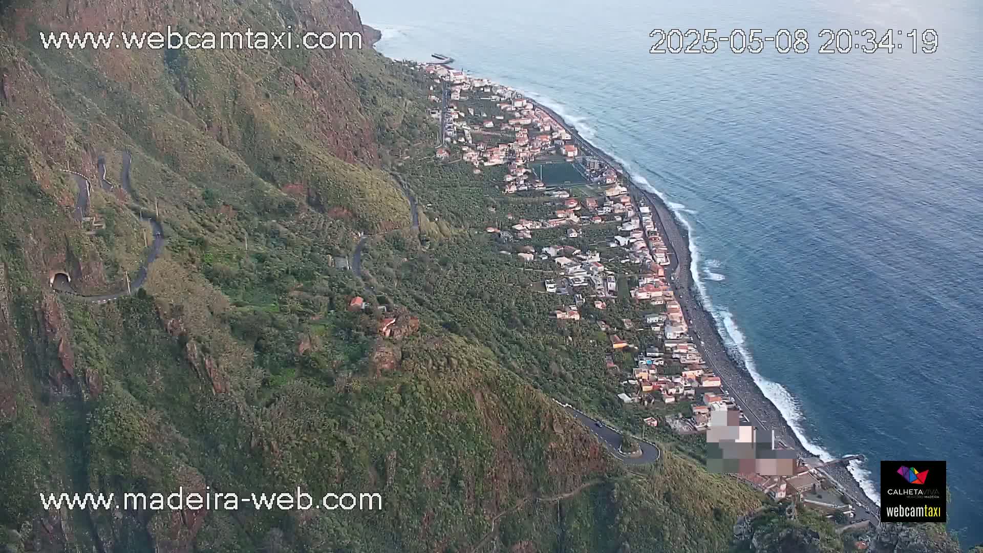 An aerial view shows a coastal town nestled between a steep, green mountain and a dark-blue ocean with gentle waves under a clear sky.
