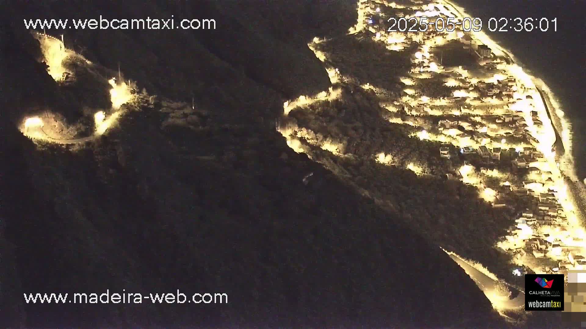 A nighttime aerial view shows a coastal town illuminated, nestled on a dark hillside with winding roads leading up to it.