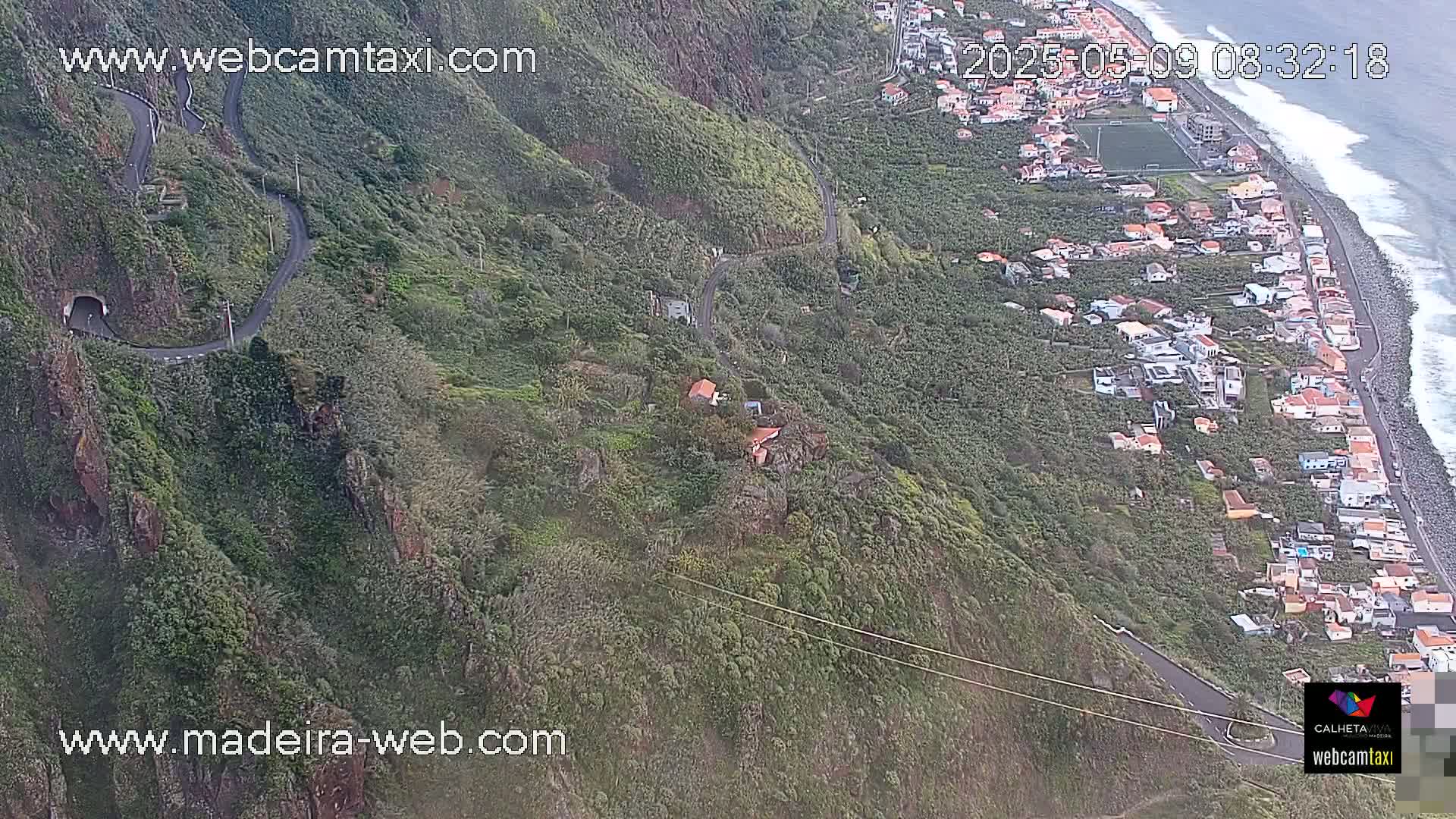 A winding road snakes down a lush green hillside toward a coastal village with a rocky shoreline and ocean waves.