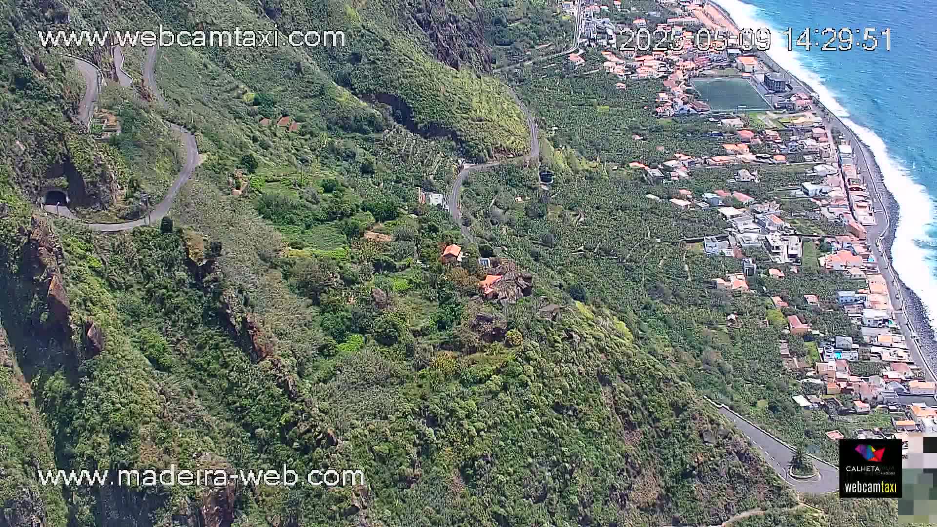 An aerial view shows a winding road along a rocky, green hillside overlooking a coastal town and ocean under sunny conditions.