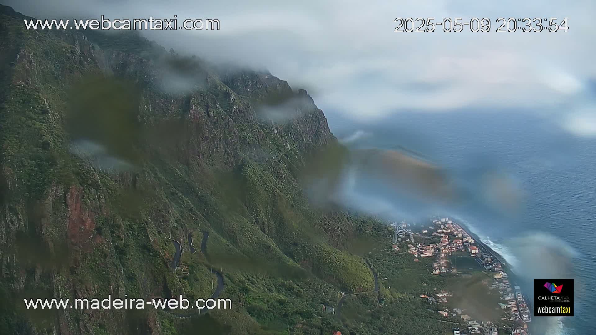 A rain-covered view shows a verdant, mountainous coastline descending to a town and ocean.