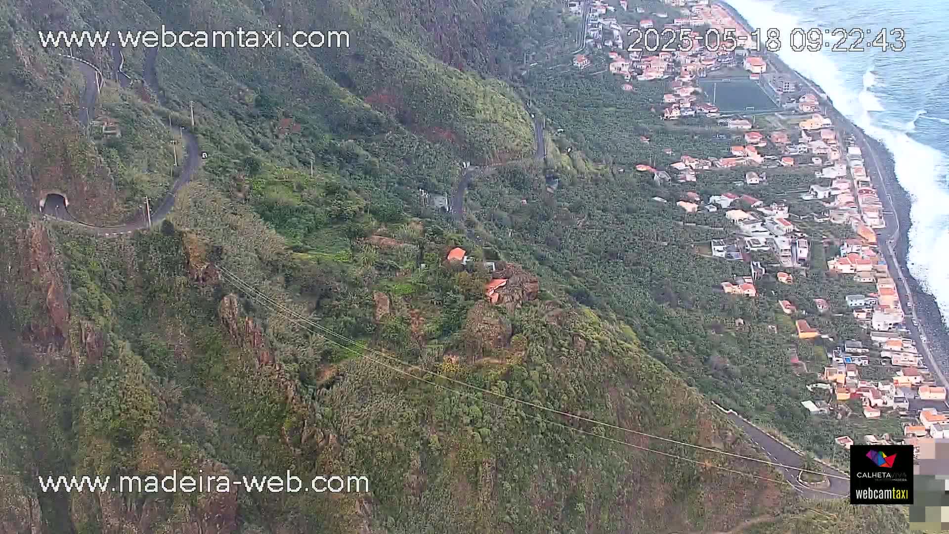 An aerial view shows a winding road carved into a lush green mountainside overlooking a coastal town and ocean with moderate waves.