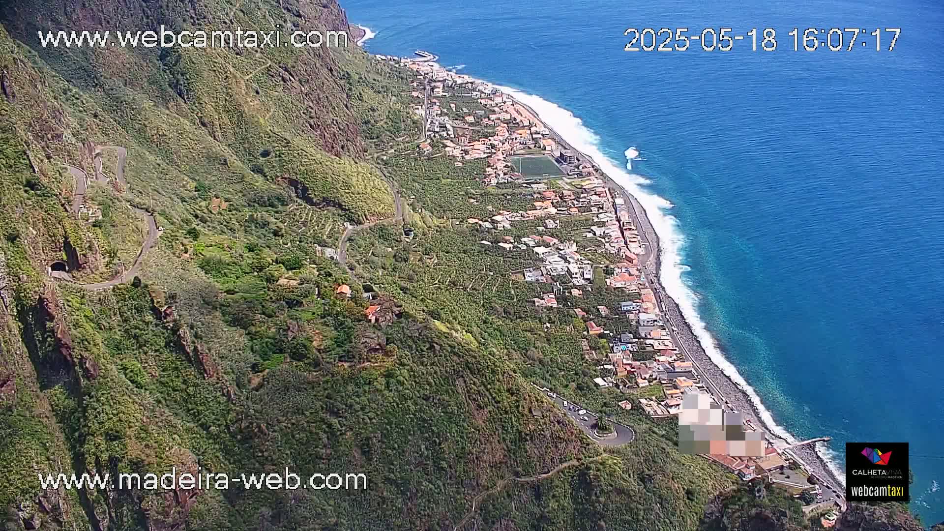A coastal town nestled between a steep, verdant mountain and a clear blue ocean with small waves is viewed from above on a sunny day.