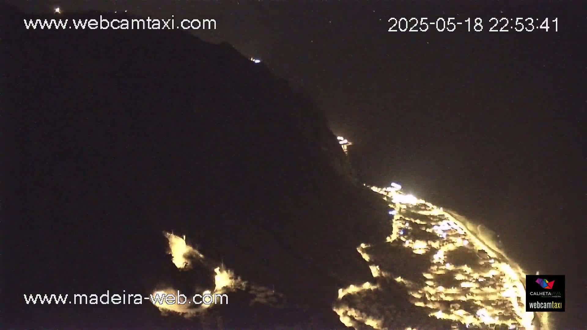A nighttime, high-angle view shows a coastal town illuminated against a dark, mountainous backdrop under a clear sky.