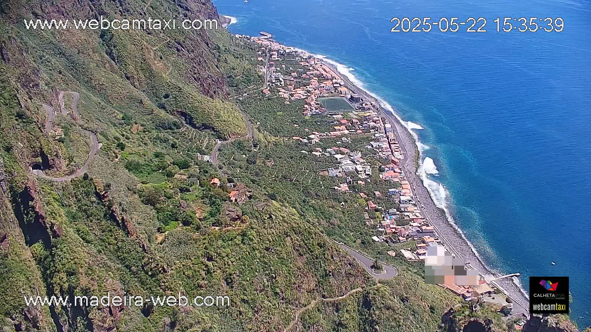 An aerial view shows a coastal town nestled between a lush green mountainside and a blue ocean with small waves on a sunny day.