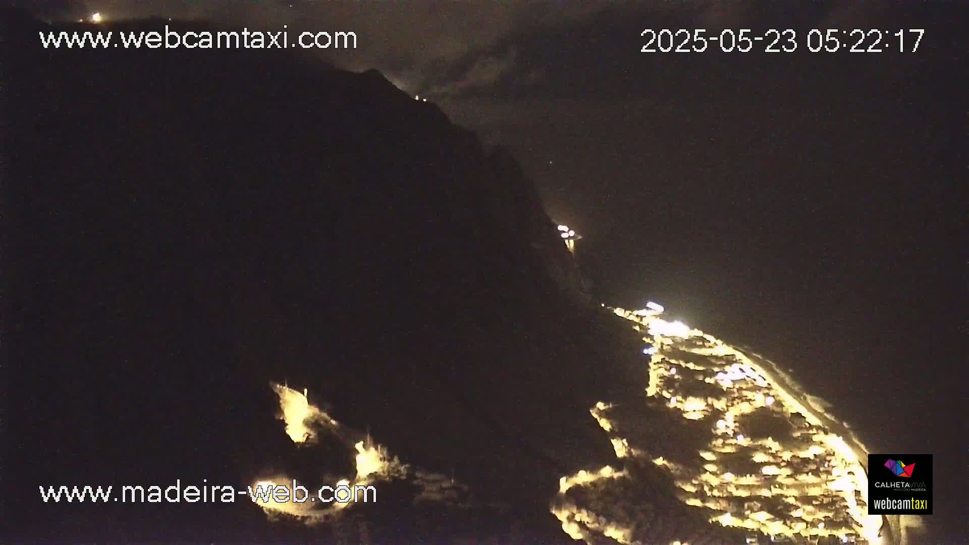 A nighttime, high-angle view shows a coastal town illuminated against a dark mountainside under a mostly clear sky.