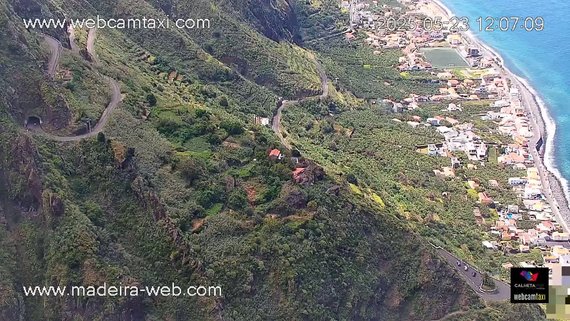 A winding road snakes through a lush, green hillside overlooking a coastal town and ocean under sunny skies.