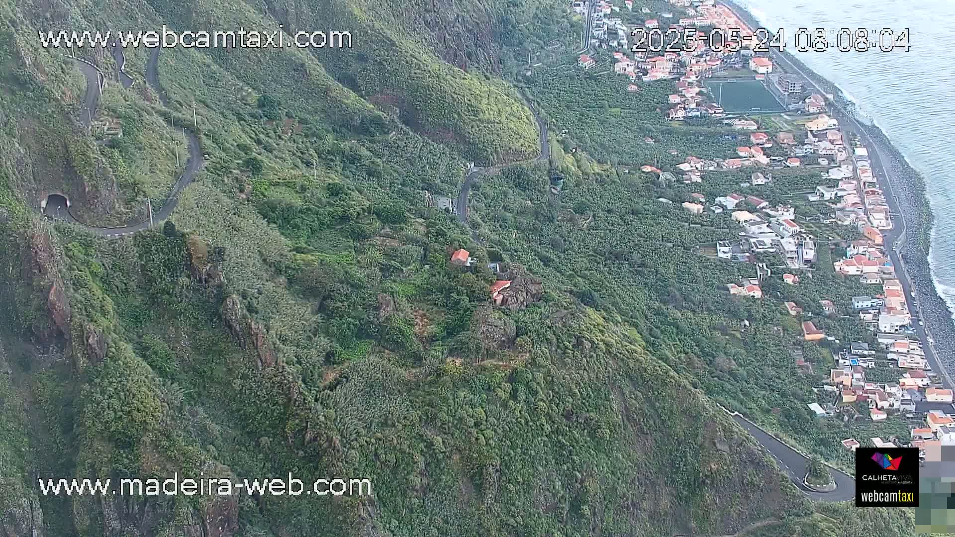 A winding road snakes through lush green hillsides overlooking a coastal town and ocean, under a partly cloudy sky.