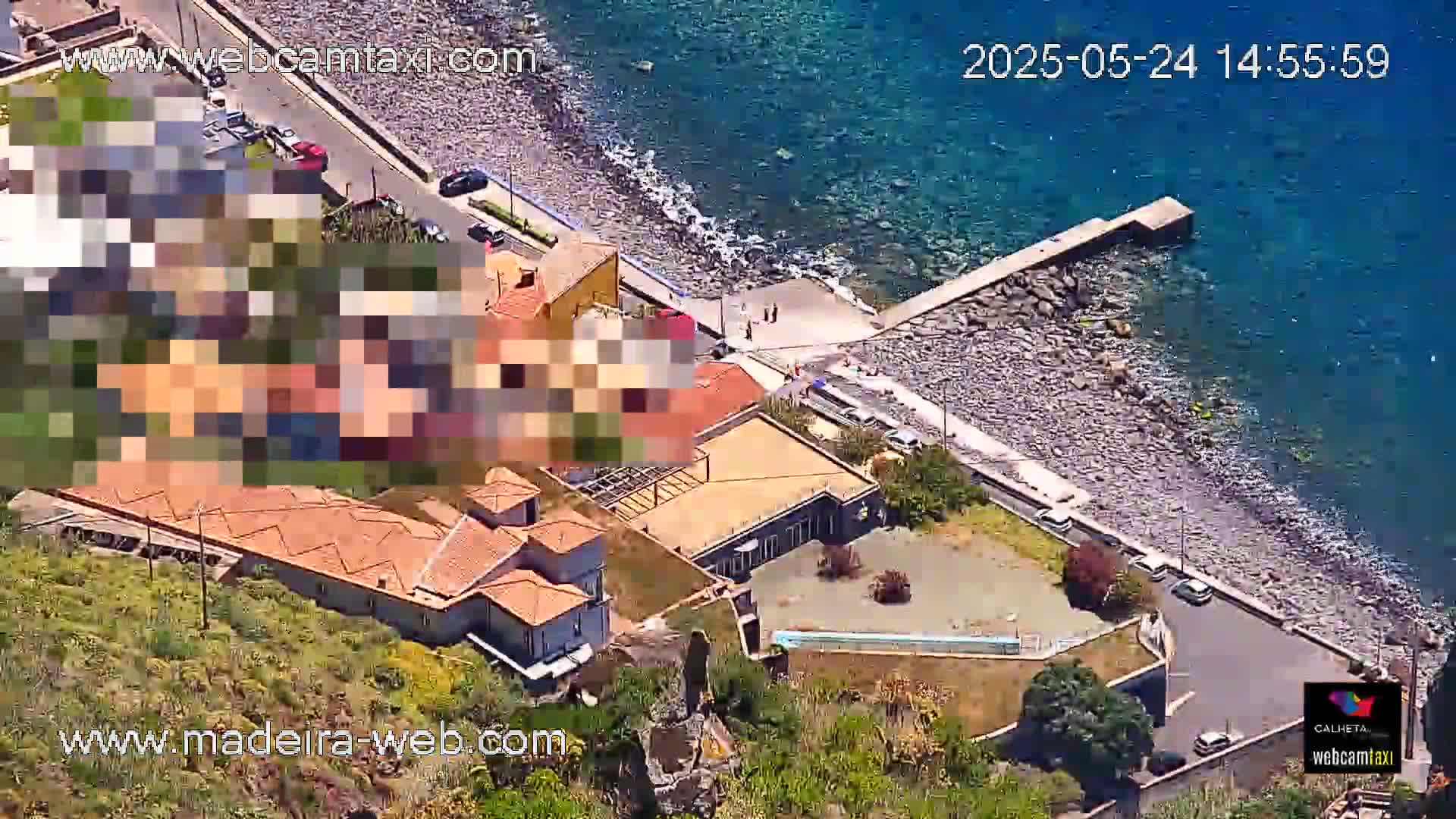 An aerial view shows a rocky coastline with calm blue water, buildings, and a small parking area.