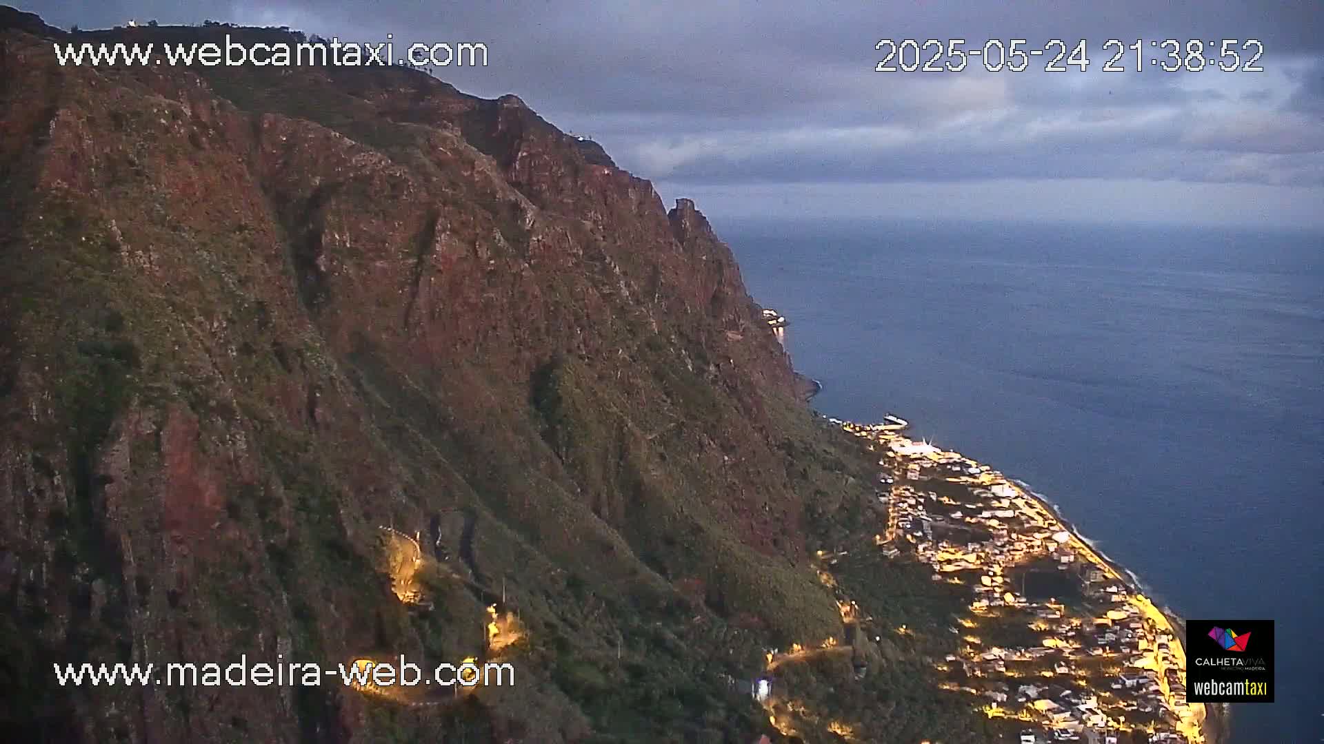 A coastal town at night is nestled at the base of a steep, dark cliff overlooking a calm, deep-blue ocean under a cloudy twilight sky.