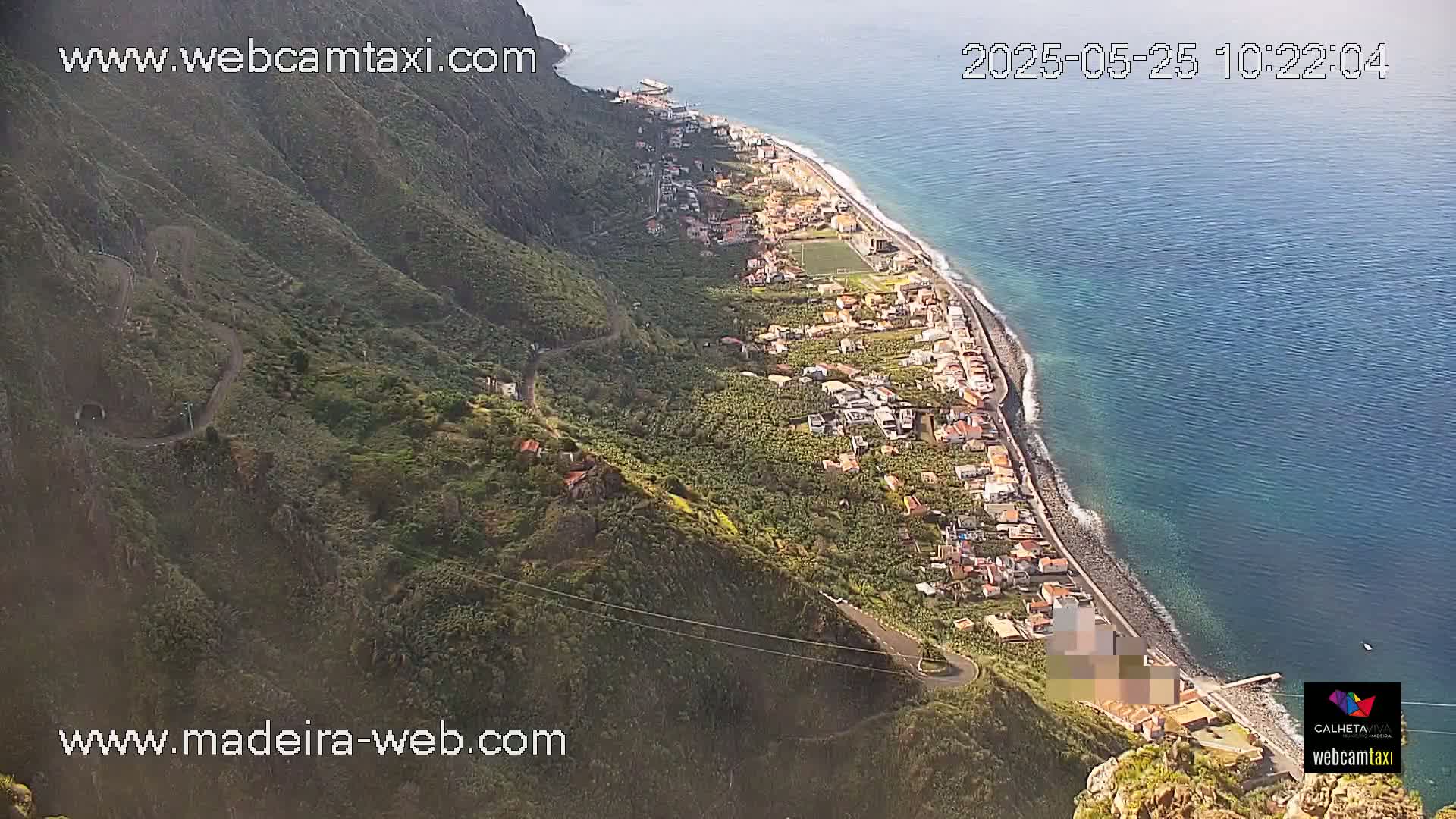 An aerial view shows a coastal town nestled between a steep, verdant mountainside and a calm, blue ocean on a sunny day.