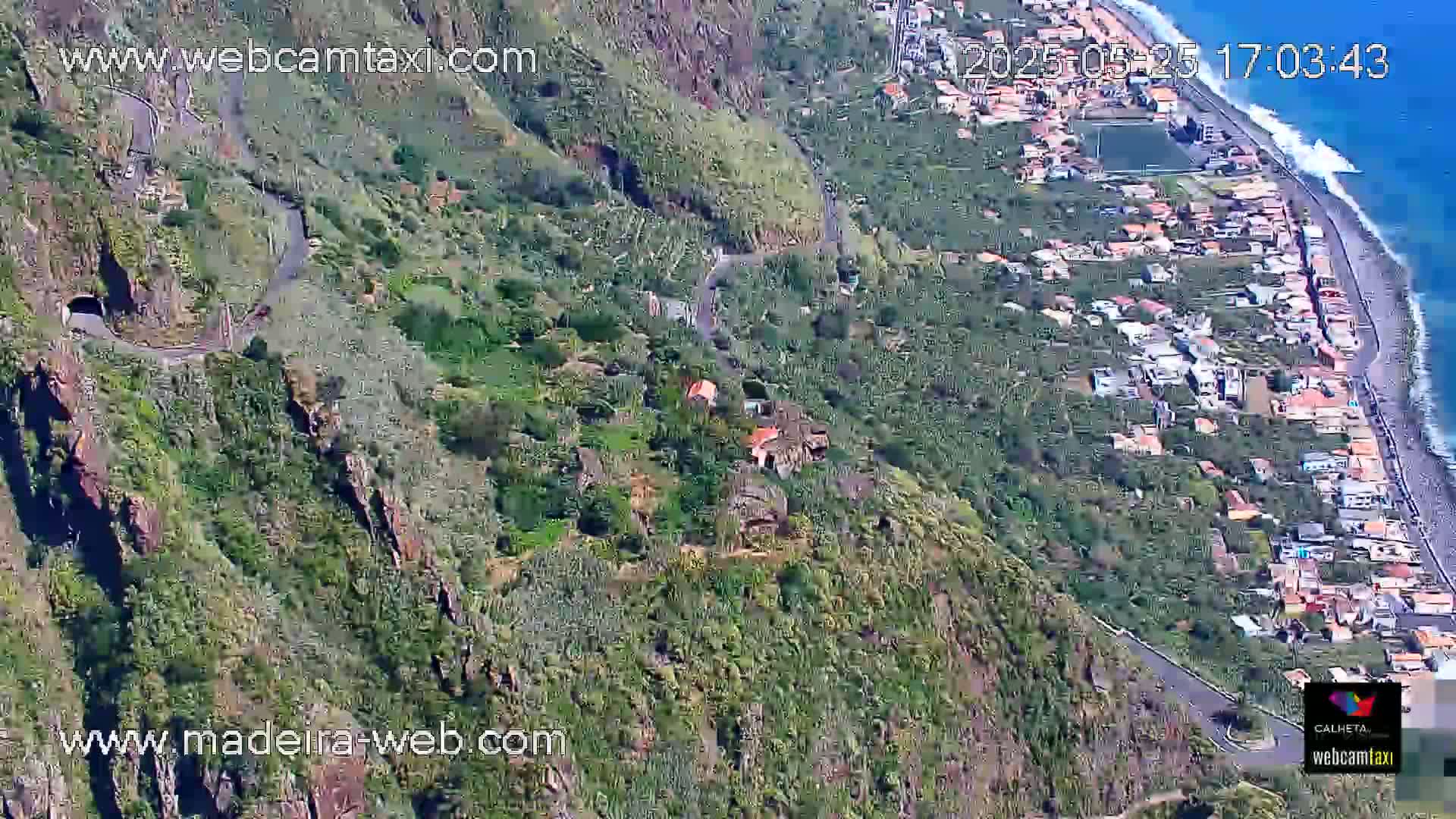 An aerial view shows a winding road descending a steep, verdant hillside toward a coastal town and ocean under sunny conditions.