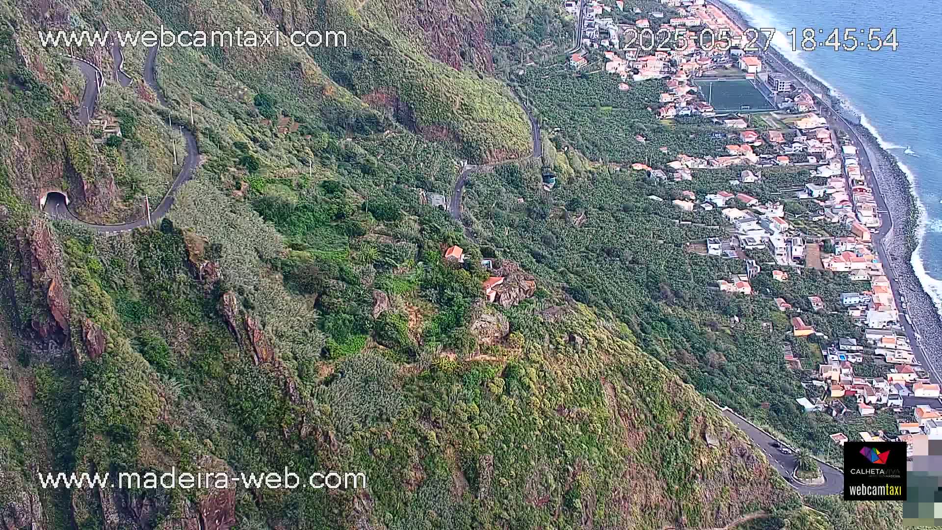 A winding road snakes down a lush, green hillside toward a coastal town with a rocky shoreline and ocean visible on a partly sunny day.