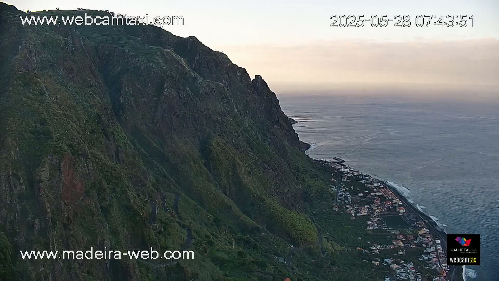 A steep, green cliff overlooking a calm ocean and a coastal town at dawn.
