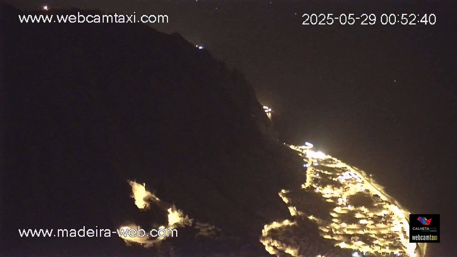 A nighttime, high-angle view shows a coastal town illuminated against a dark mountainside under a clear, starry sky.