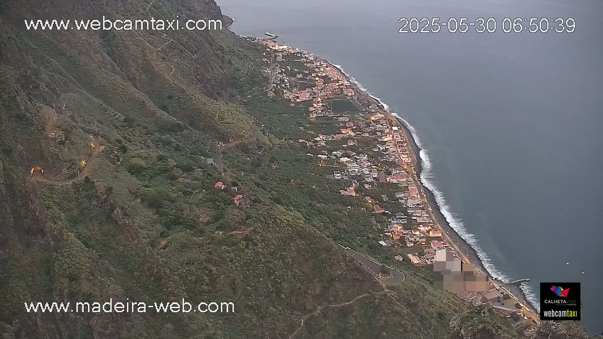 An aerial view shows a coastal town nestled between a dark green mountainside and a dark blue ocean under an overcast sky.
