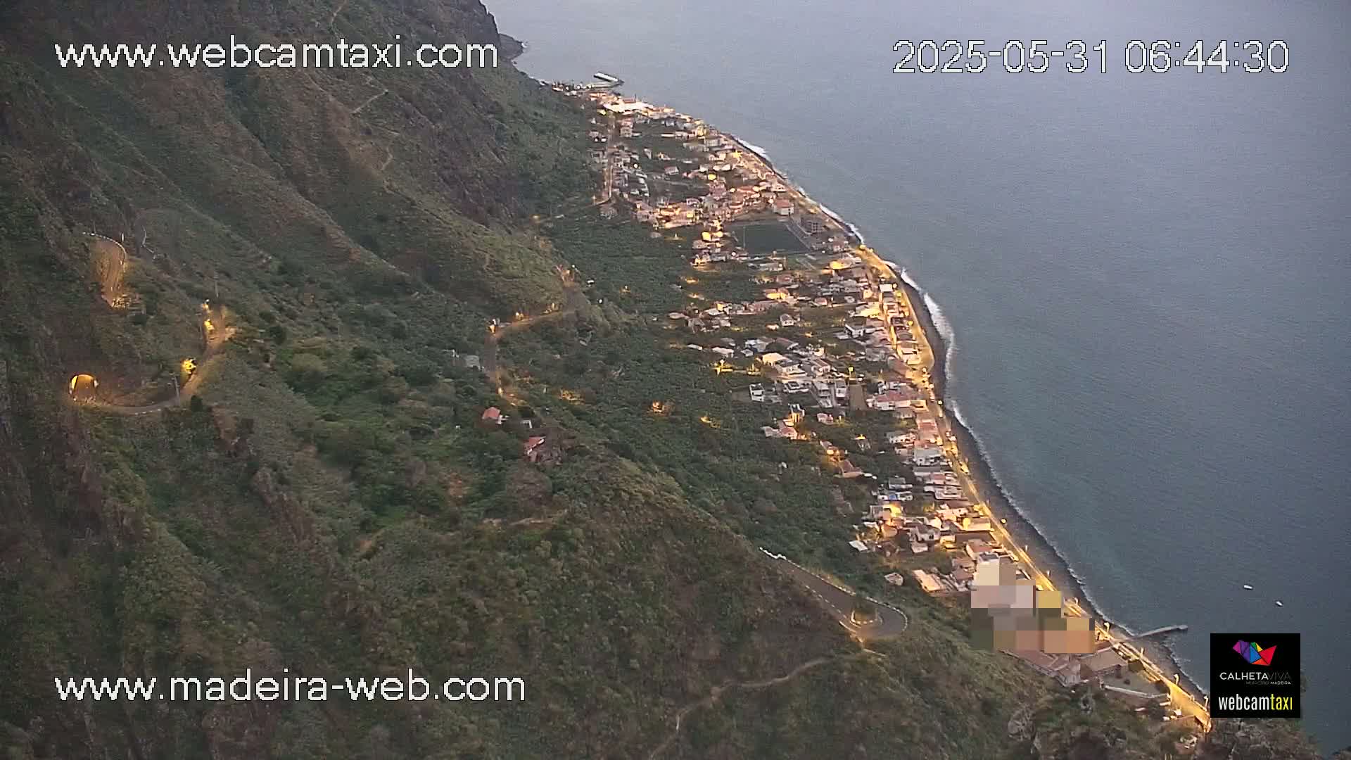 A coastal town nestled between a dark green mountain and a calm, dark blue ocean at twilight.