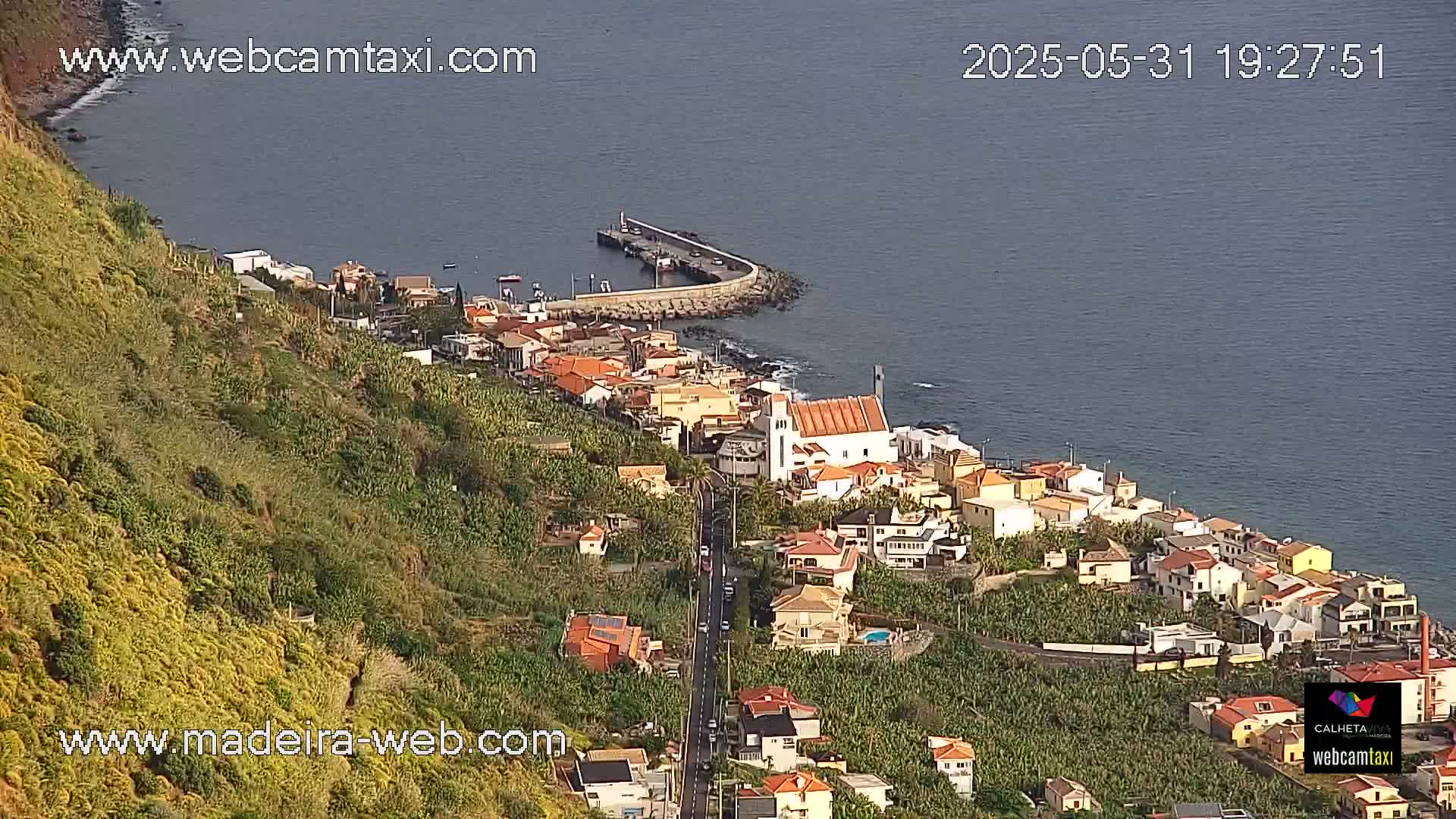 An aerial view shows a coastal town nestled on a hillside overlooking a calm ocean on a sunny day.