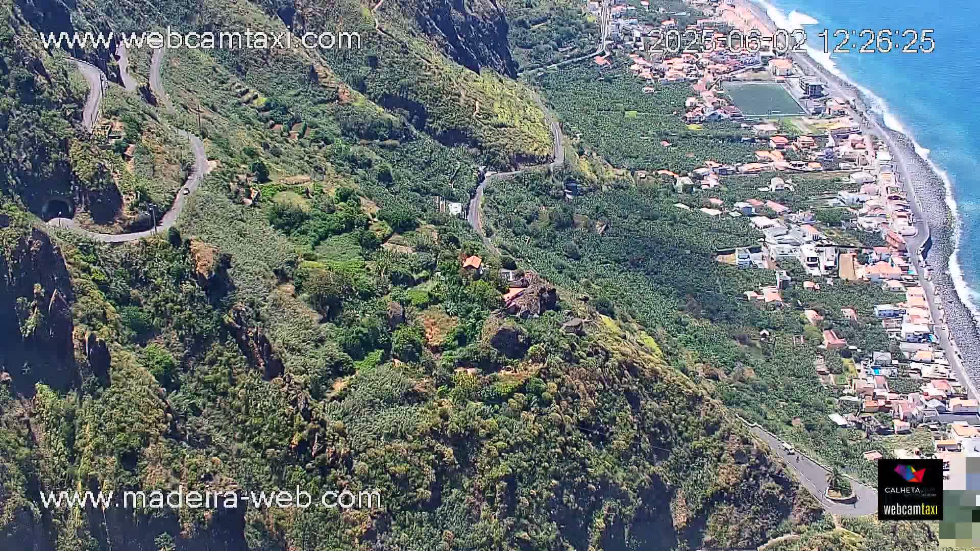 A winding road follows a cliffside overlooking a coastal town and ocean under sunny conditions.