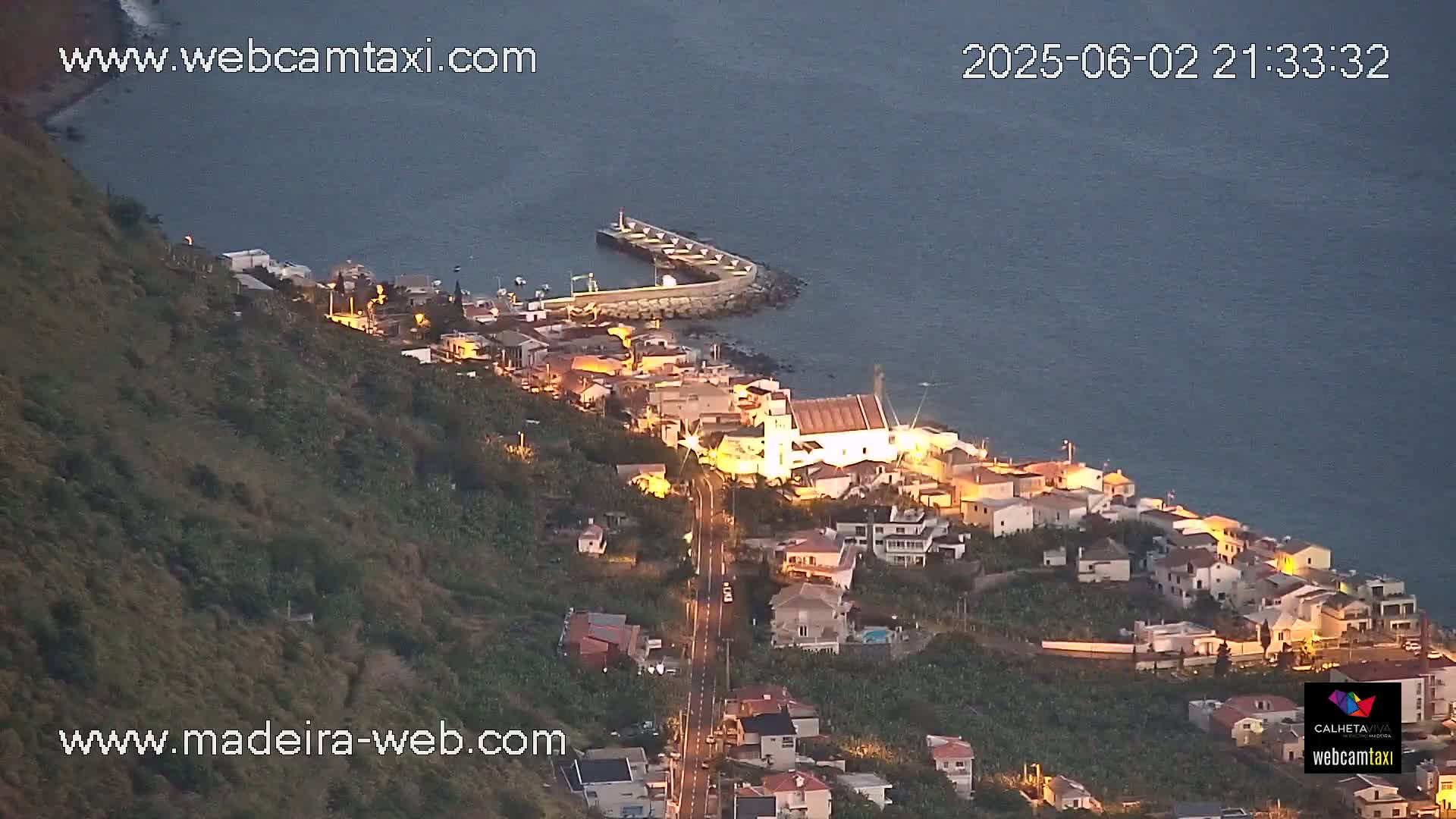 A coastal town with illuminated buildings is nestled on a hillside overlooking a calm, dark-blue sea at twilight.