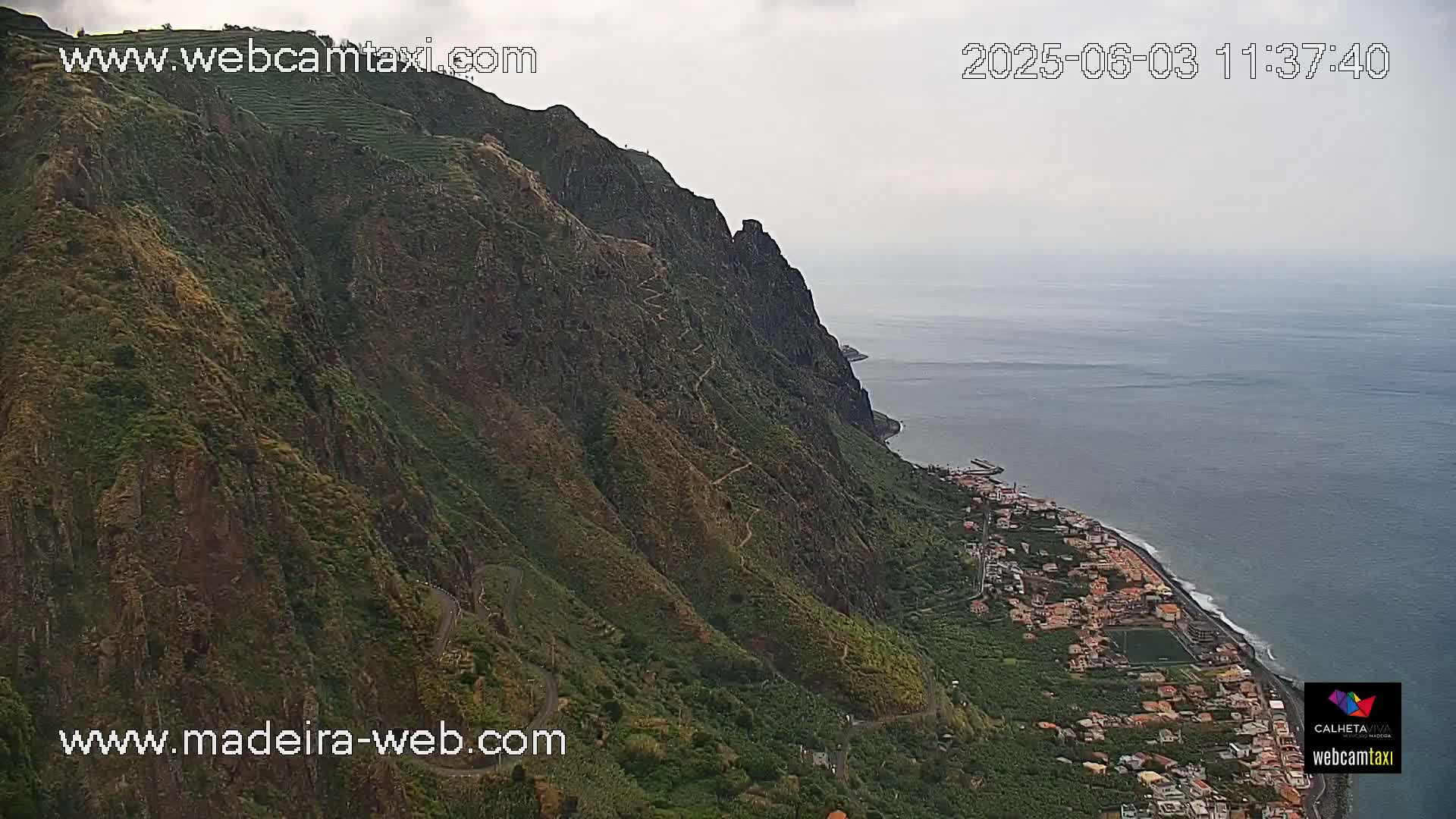 A winding road follows a steep, green, mountainous coastline that drops sharply to a calm ocean under an overcast sky.