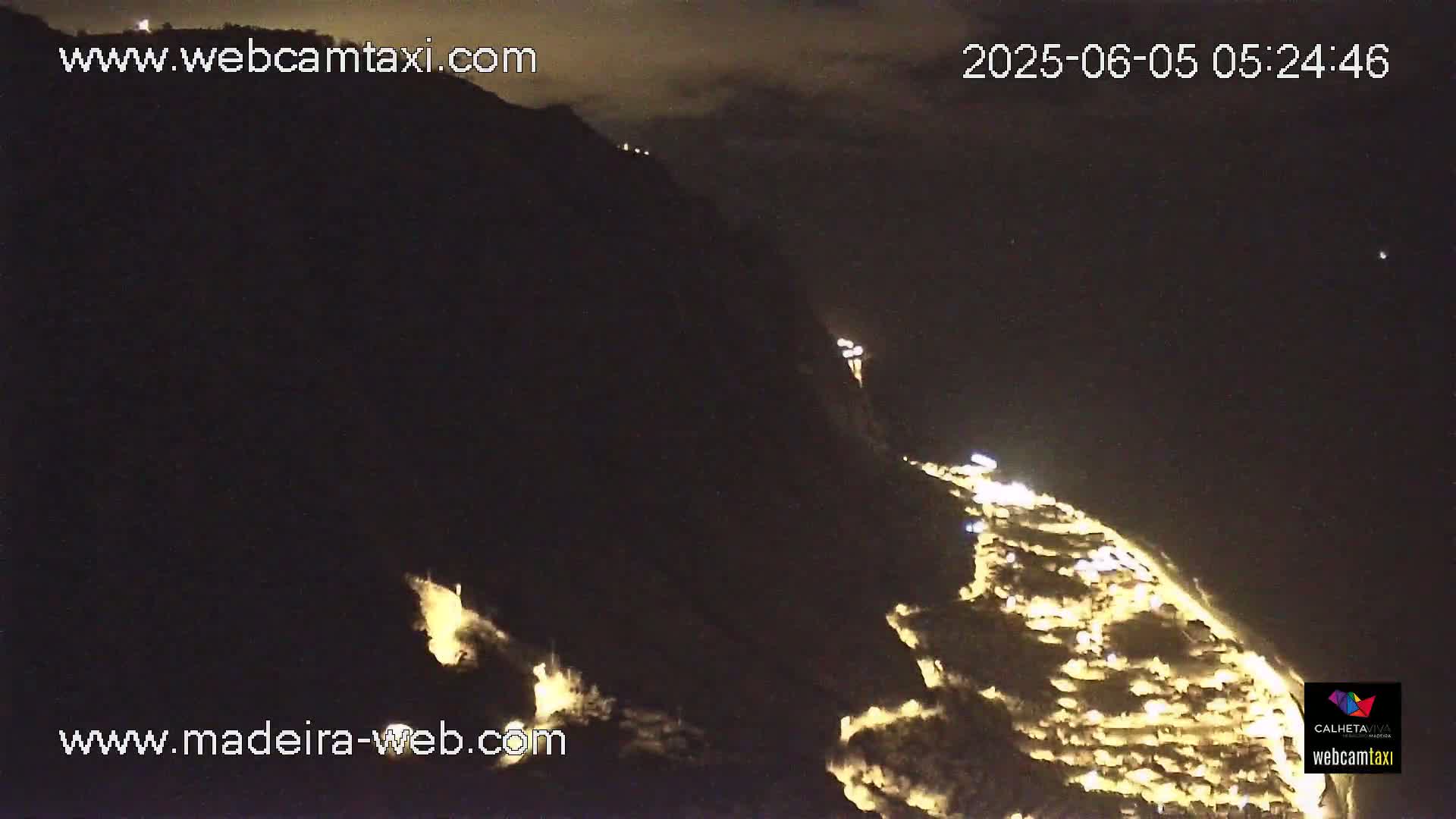 A nighttime long shot shows a hillside town illuminated with lights, nestled below a dark mountain under a partly cloudy sky.