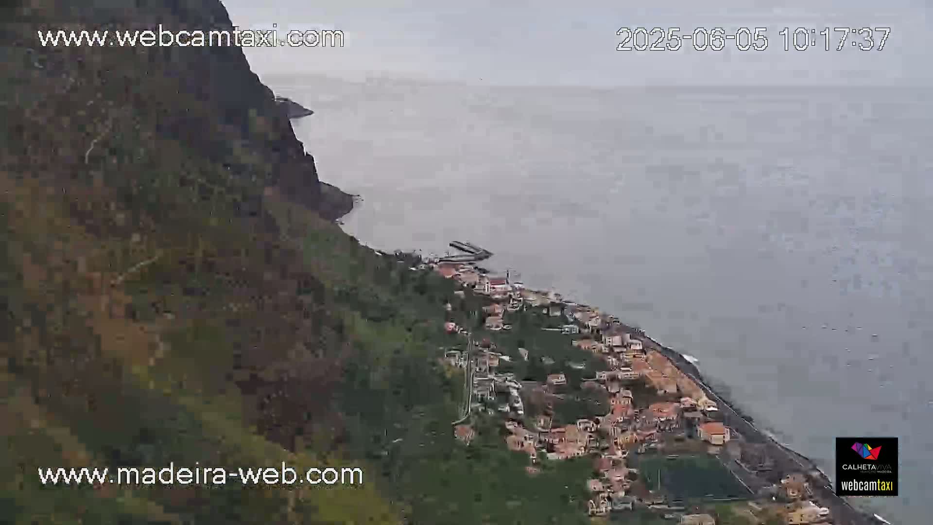 A coastal town nestled against a steep, dark cliff overlooking a calm, gray ocean on an overcast day.