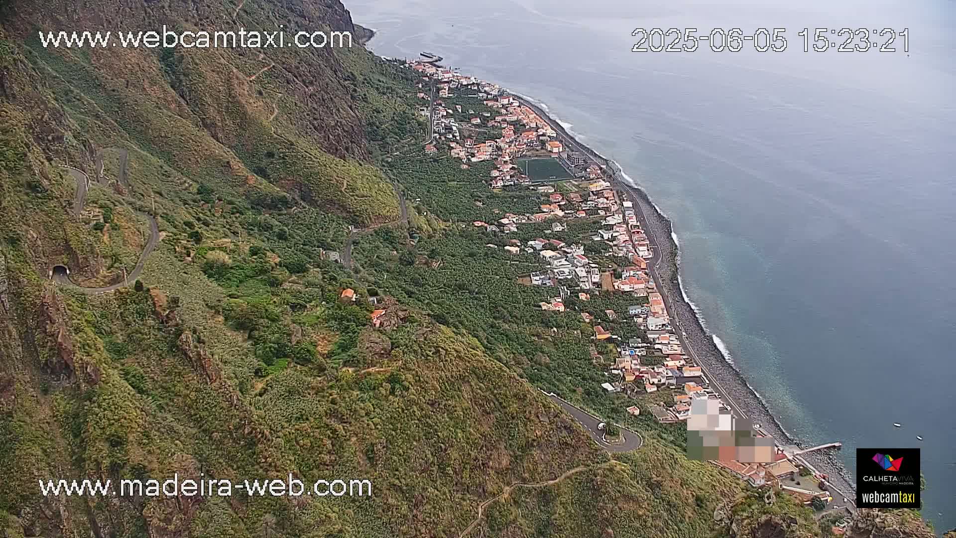 An aerial view shows a coastal town nestled between a steep, green mountainside and a dark blue ocean under somewhat overcast skies.