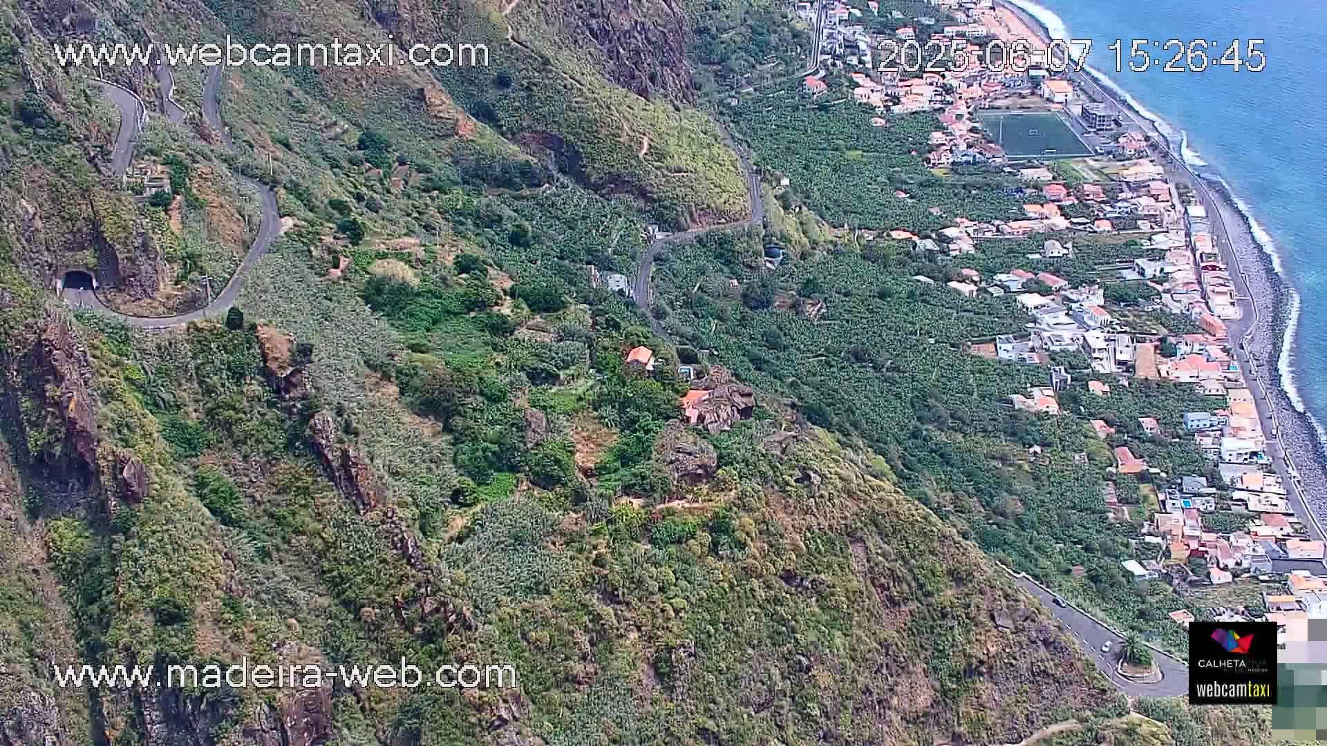 A winding road snakes down a lush, green hillside toward a coastal town and ocean under a clear sky.