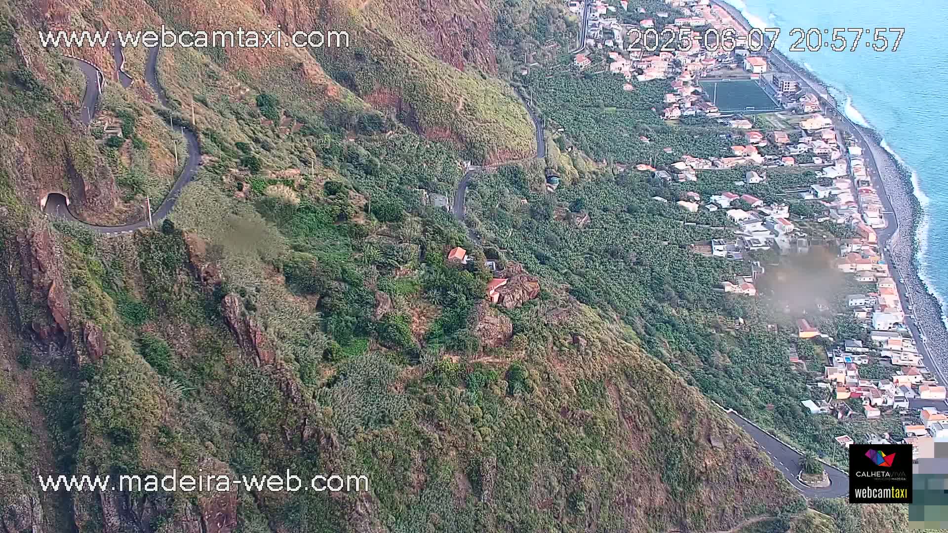 A winding road snakes down a steep, verdant hillside overlooking a coastal town and ocean under a partly cloudy sky.