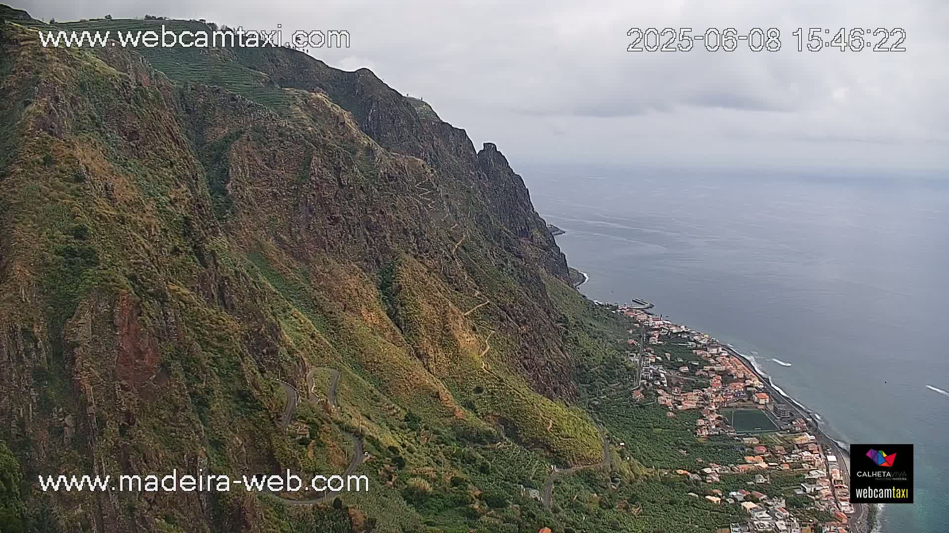 A winding road descends a steep, verdant cliffside overlooking a coastal town and the ocean under an overcast sky.
