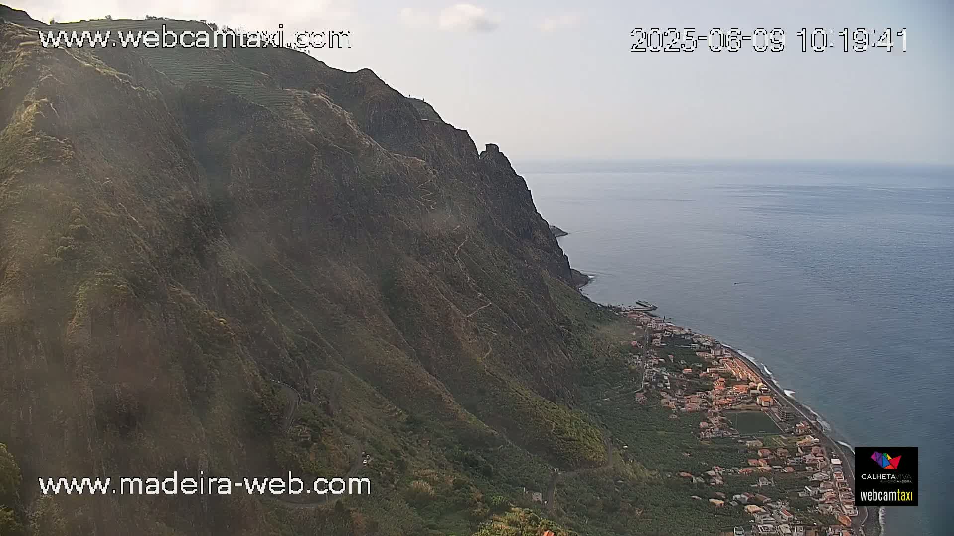 A steep, verdant cliff overlooks a calm ocean and a coastal town on a partly cloudy day.