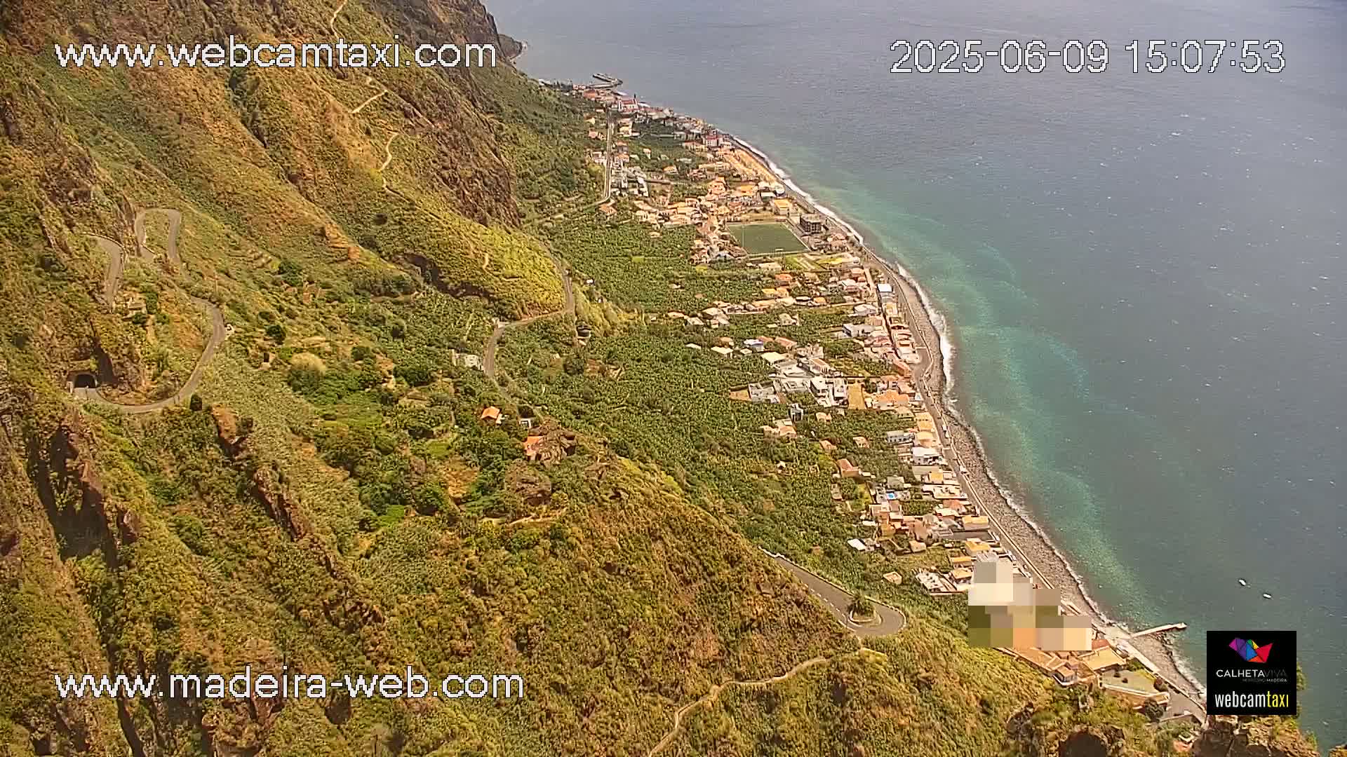 An aerial view shows a coastal town nestled between a steep, winding mountain road and a clear, turquoise ocean under sunny conditions.