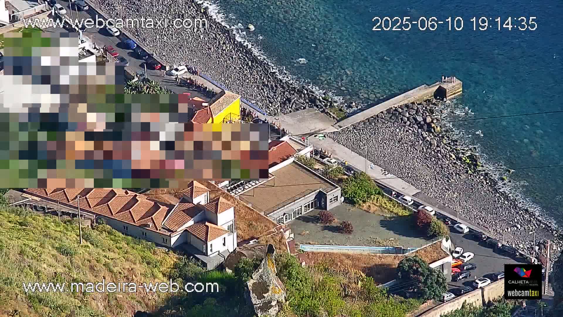 An aerial view shows a rocky coastline with a small pier, buildings, and parked cars, under sunny conditions.