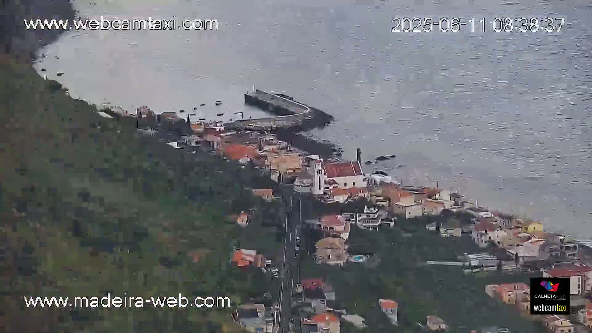An aerial view shows a coastal town with a small harbor on a partly cloudy day.