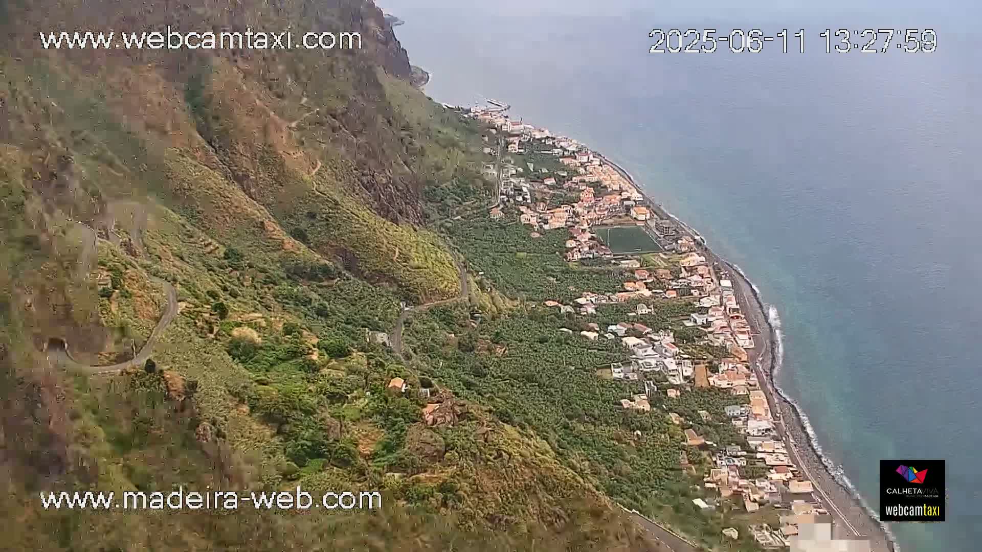 An aerial view shows a coastal town nestled between a steep, green mountain and a calm, blue ocean on a partly cloudy day.