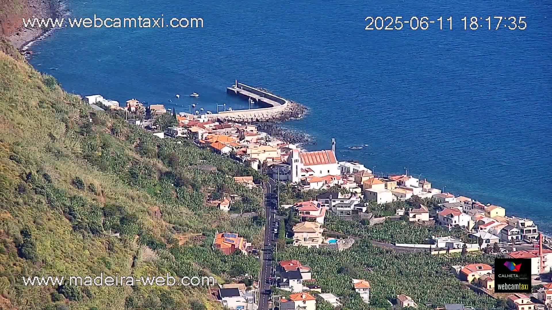 A coastal town with a church and harbor sits on a hillside overlooking a calm blue ocean under sunny skies.