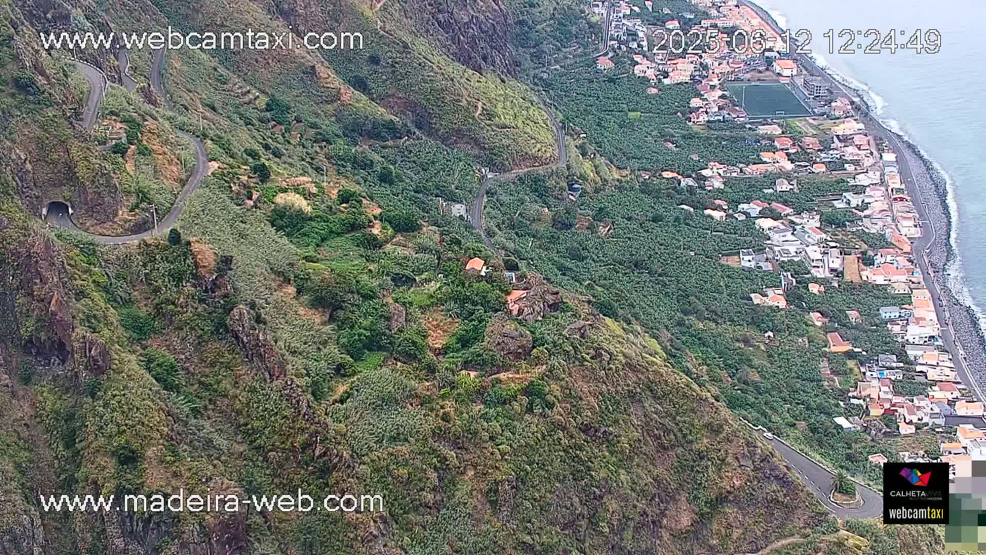 A winding road snakes down a lush, green hillside toward a coastal town and ocean, under a partly cloudy sky.