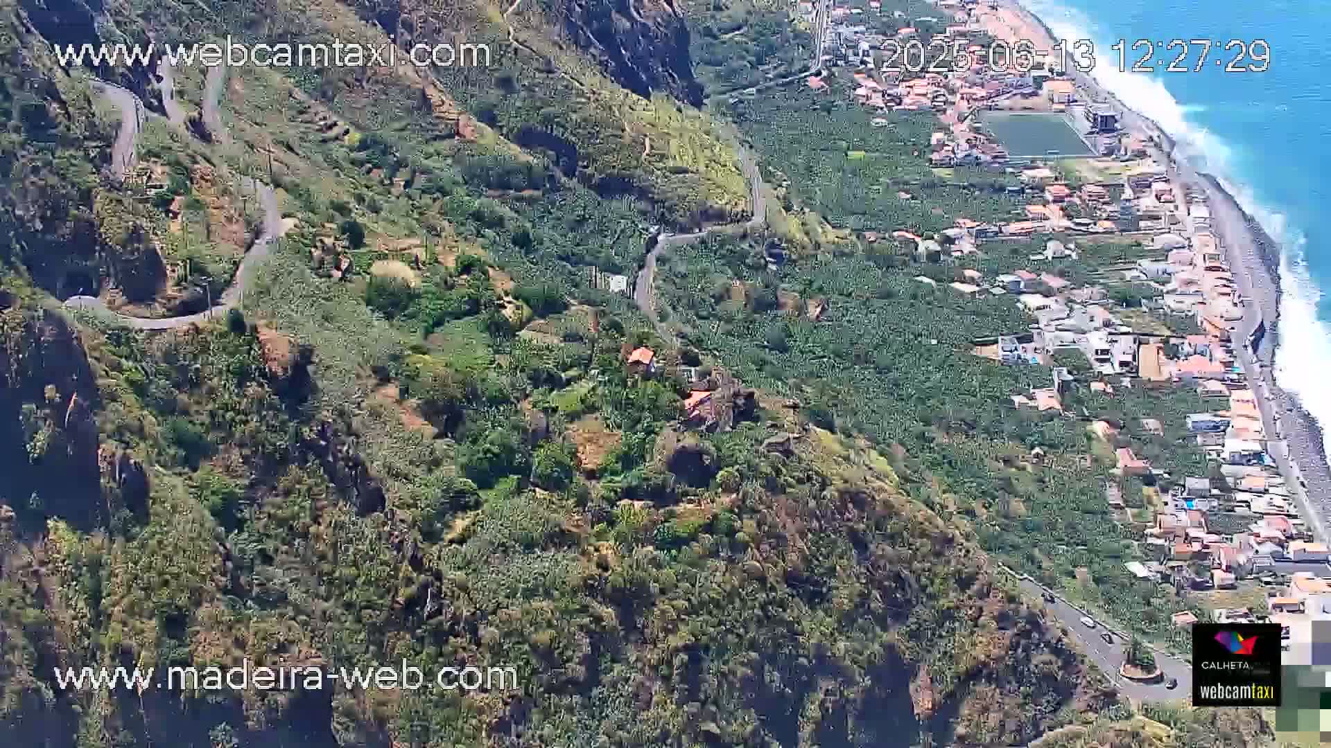 An aerial view shows a winding road on a steep, green hillside overlooking a coastal town and ocean with calm waves.