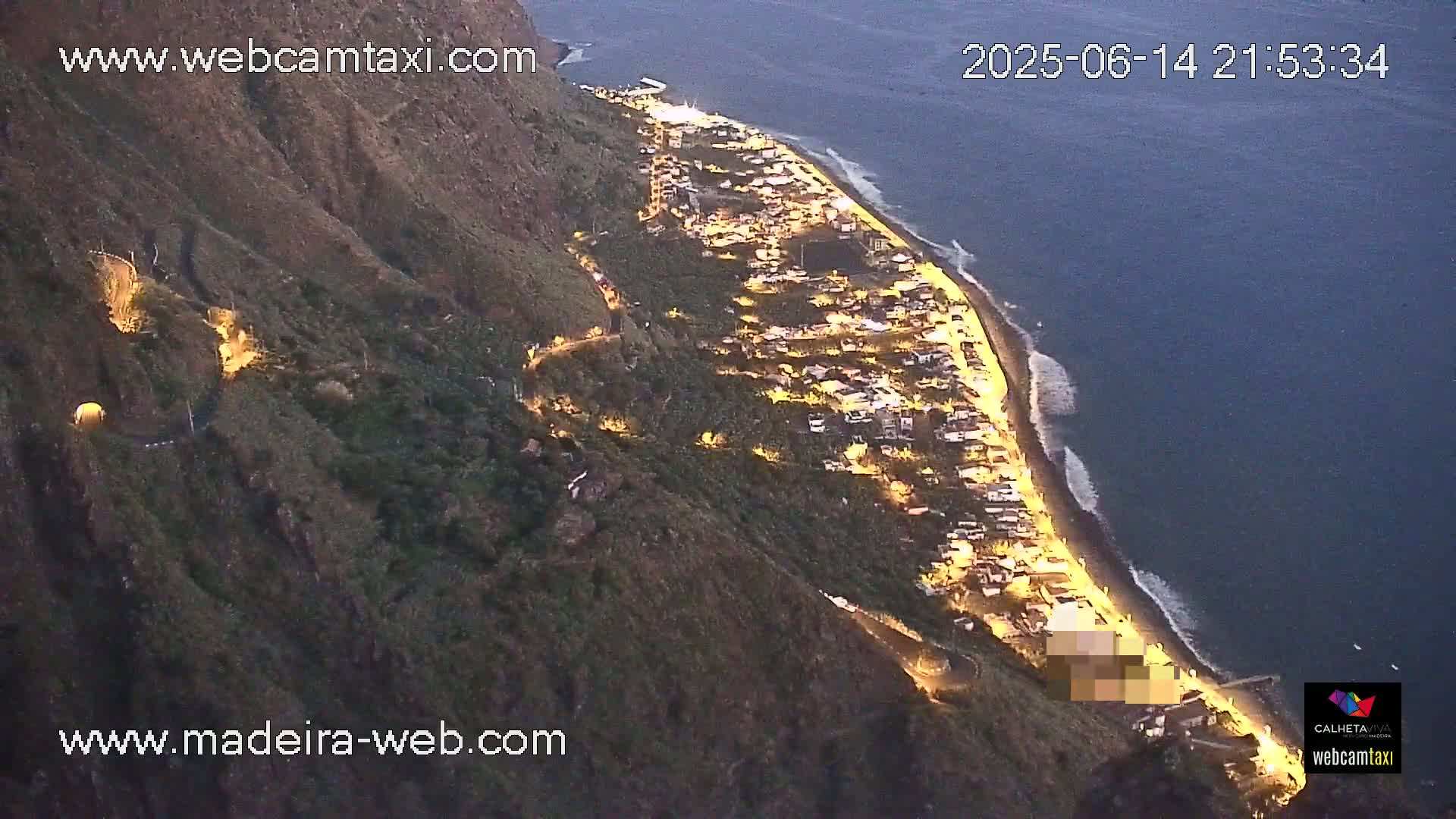 An aerial view shows a coastal town nestled on a hillside overlooking a calm ocean on a sunny day, with a small harbor and banana plantations visible.