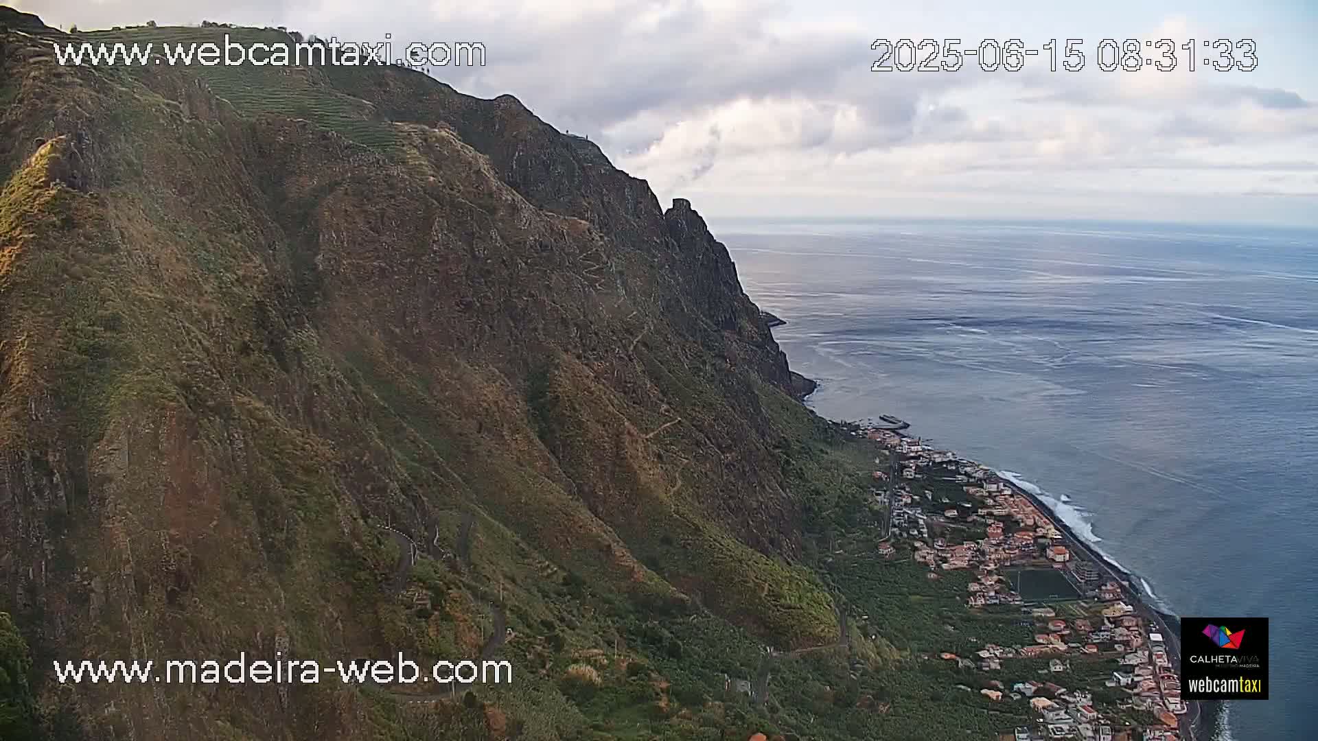 A steep, green cliffside descends to a coastal town on a partly cloudy day, with a calm ocean stretching to the horizon.