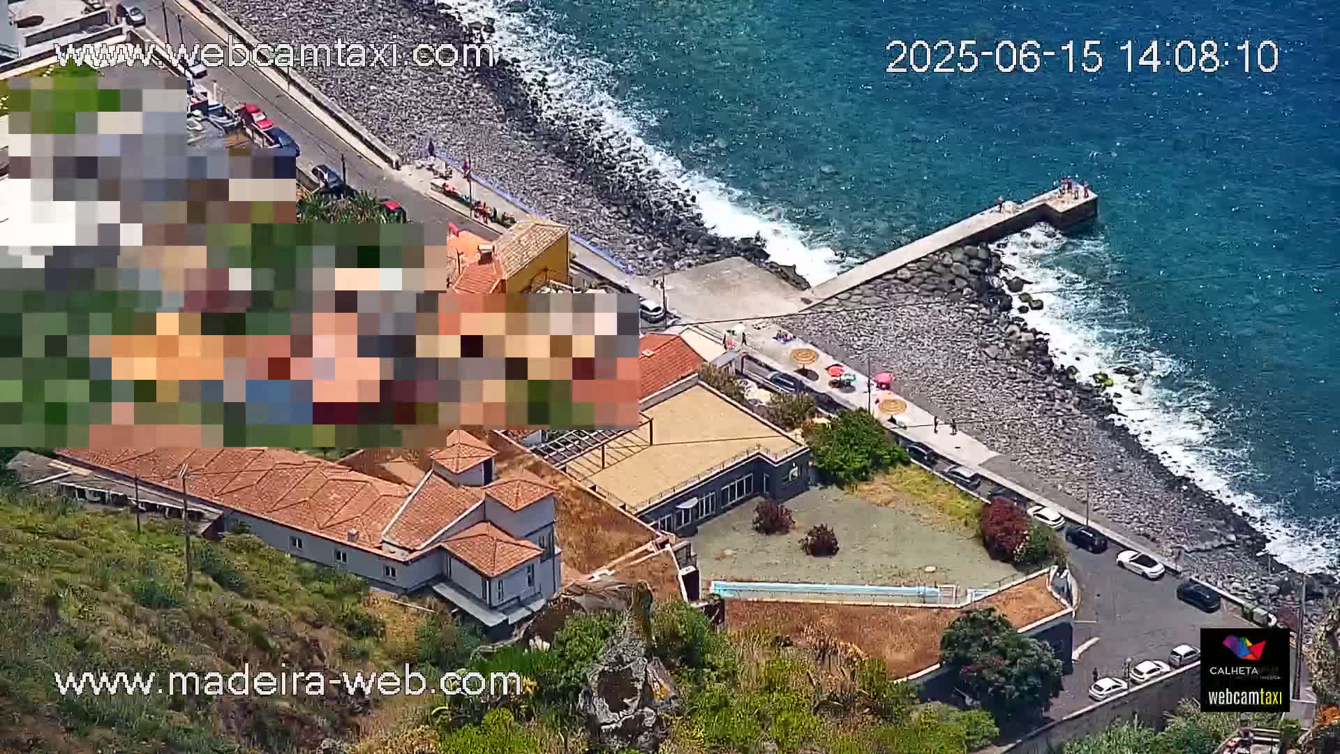An aerial view shows a coastal area with buildings, a small beach, a rocky shoreline, a pier with people on it, and a calm ocean under a sunny sky.