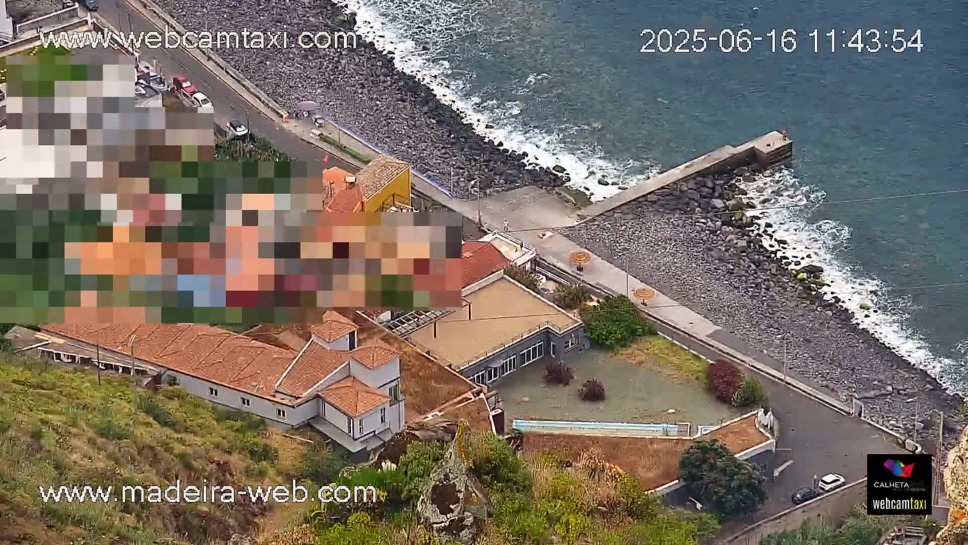 An aerial view shows a coastal area with buildings, a rocky shoreline, and a partly cloudy sky with a calm ocean.