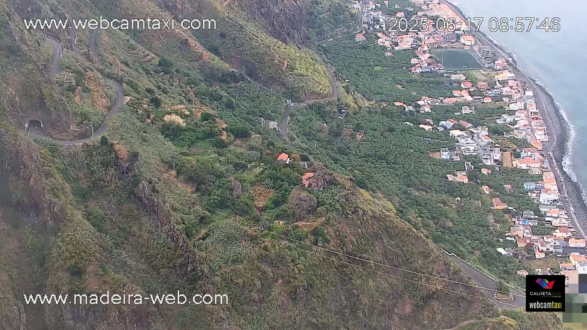 An aerial view shows a winding road along a steep, green hillside descending to a coastal town with a rocky beach and ocean under mostly cloudy skies.