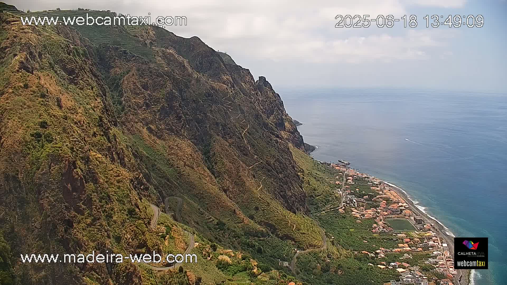 A winding road follows a steep, green cliffside overlooking a calm ocean and a coastal town under a partly cloudy sky.