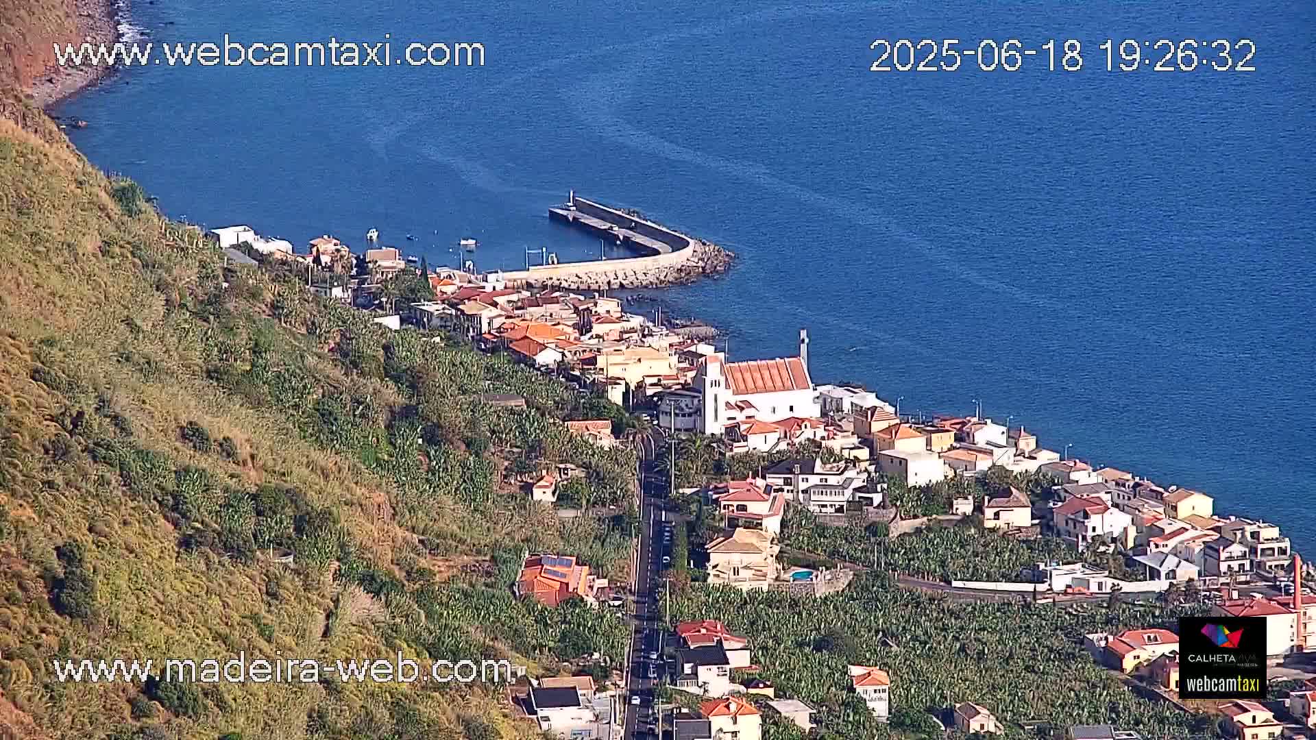 A coastal town with white-washed buildings and a church is nestled on a hillside overlooking a calm blue sea on a sunny day.
