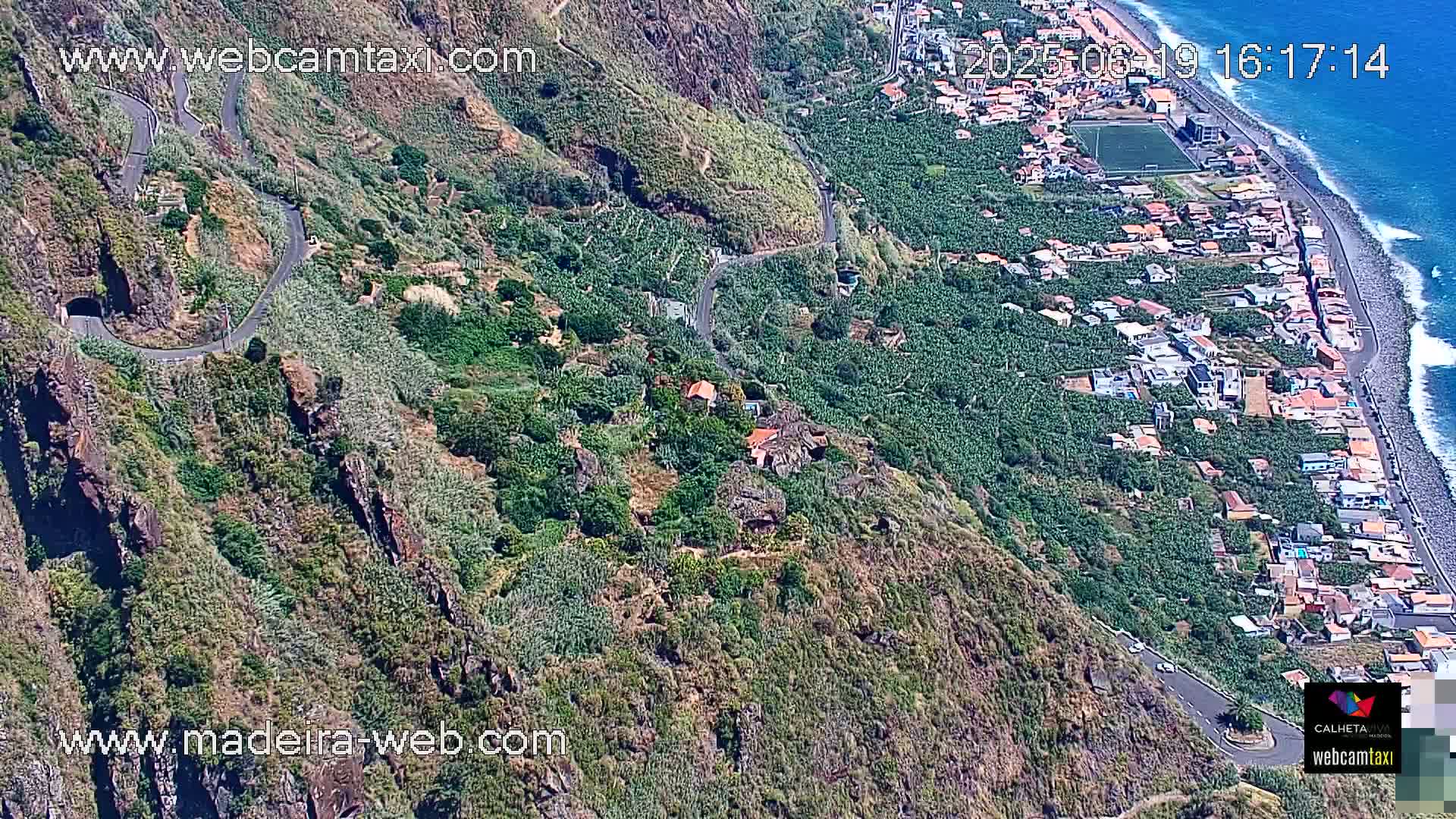 A winding road snakes down a steep, verdant hillside toward a coastal town and ocean under sunny skies.